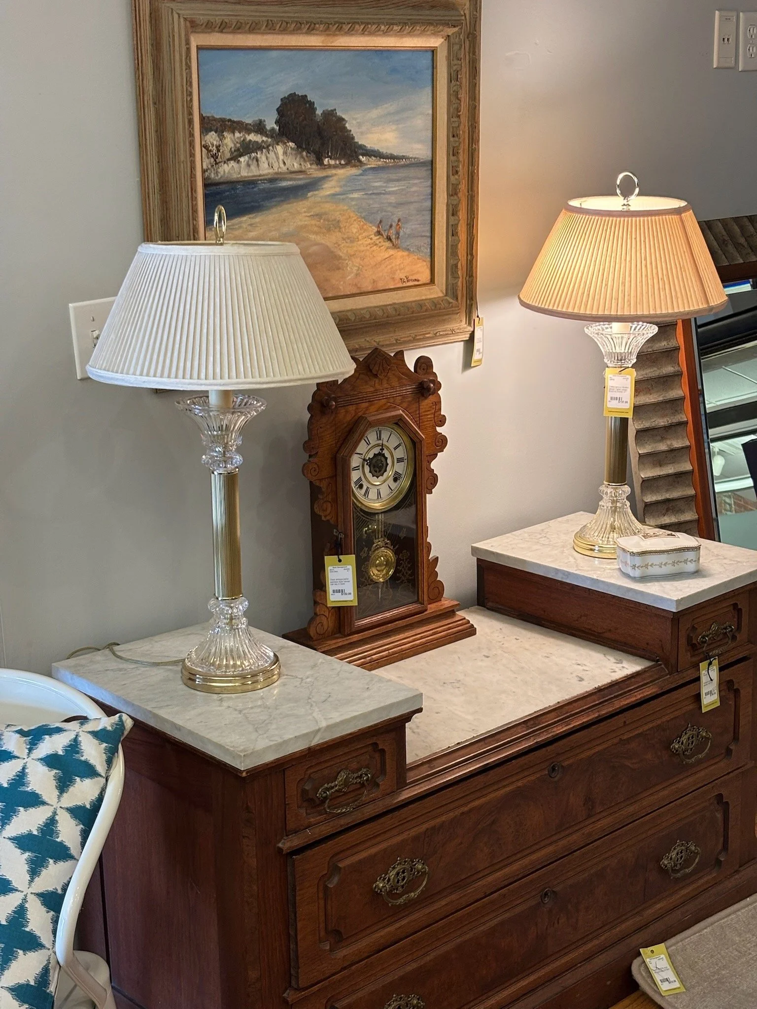 A vintage wooden dresser with a marble top, two table lamps with pleated shades, a decorative clock, and a framed landscape painting on the wall behind it.