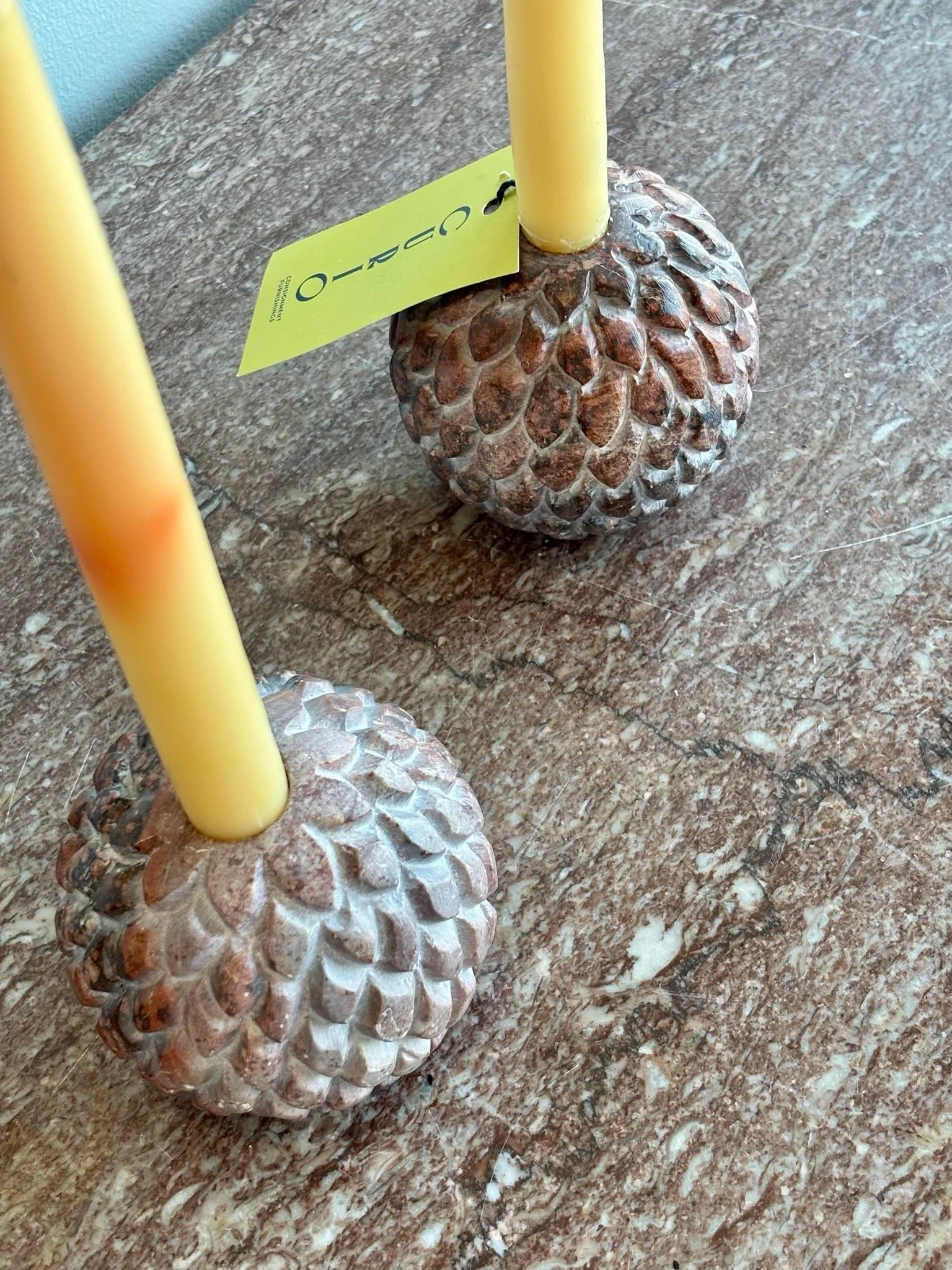 Close-up of a decorative base resembling pinecones with yellow candles inserted in the middle, placed on a speckled brown countertop. A yellow tag with black writing is attached to one of the candles.