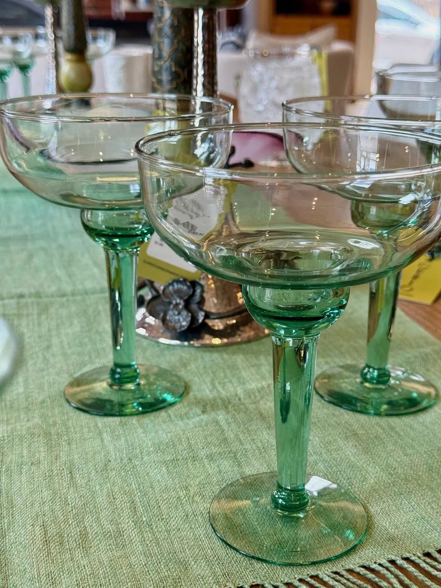 Four green glass coupe glasses placed on a green tablecloth, with various decorative items in the blurred background.