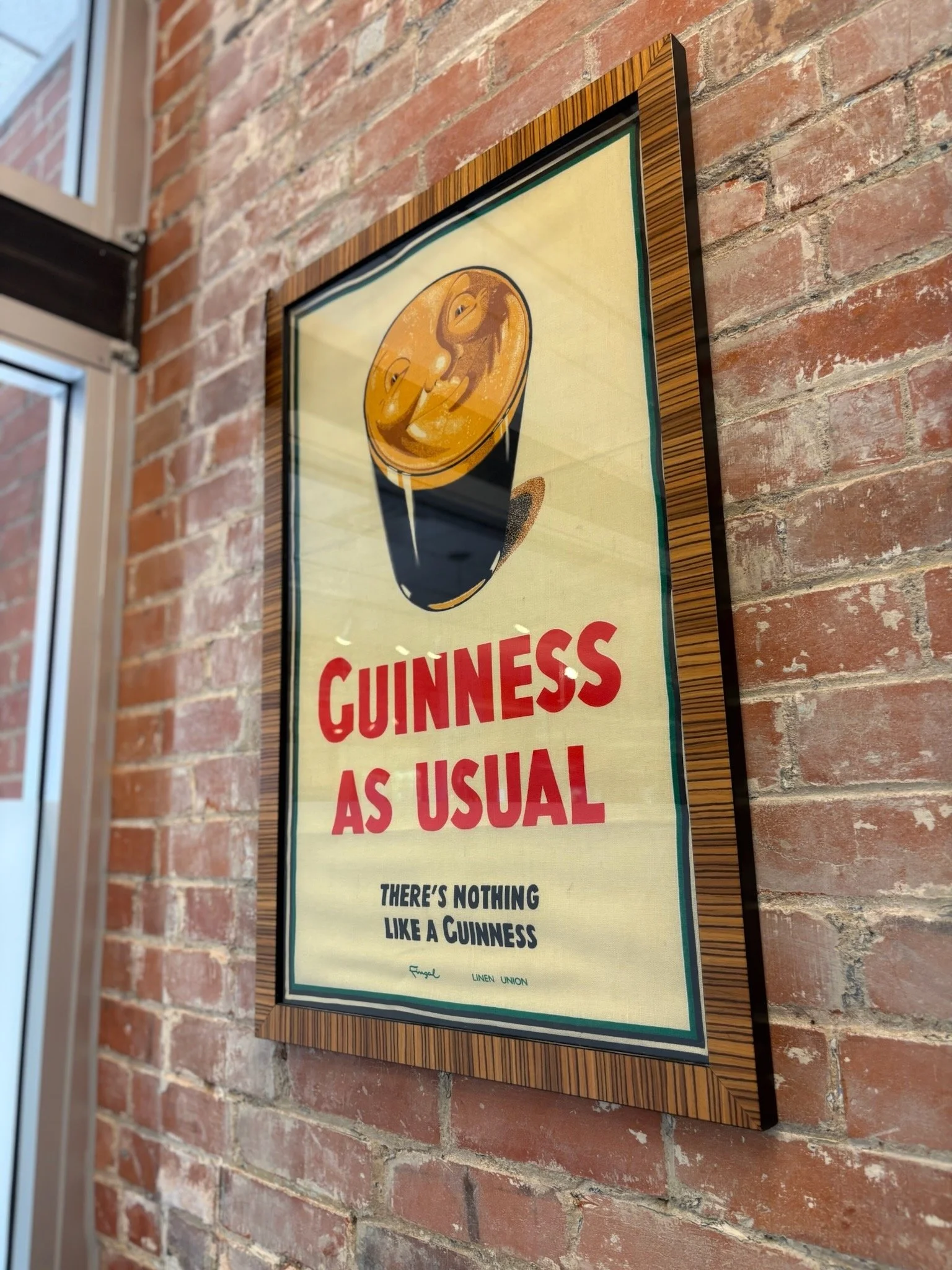 A vintage Guinness advertisement poster displaying a pint of Guinness with foam, hung on a red brick wall near a window.