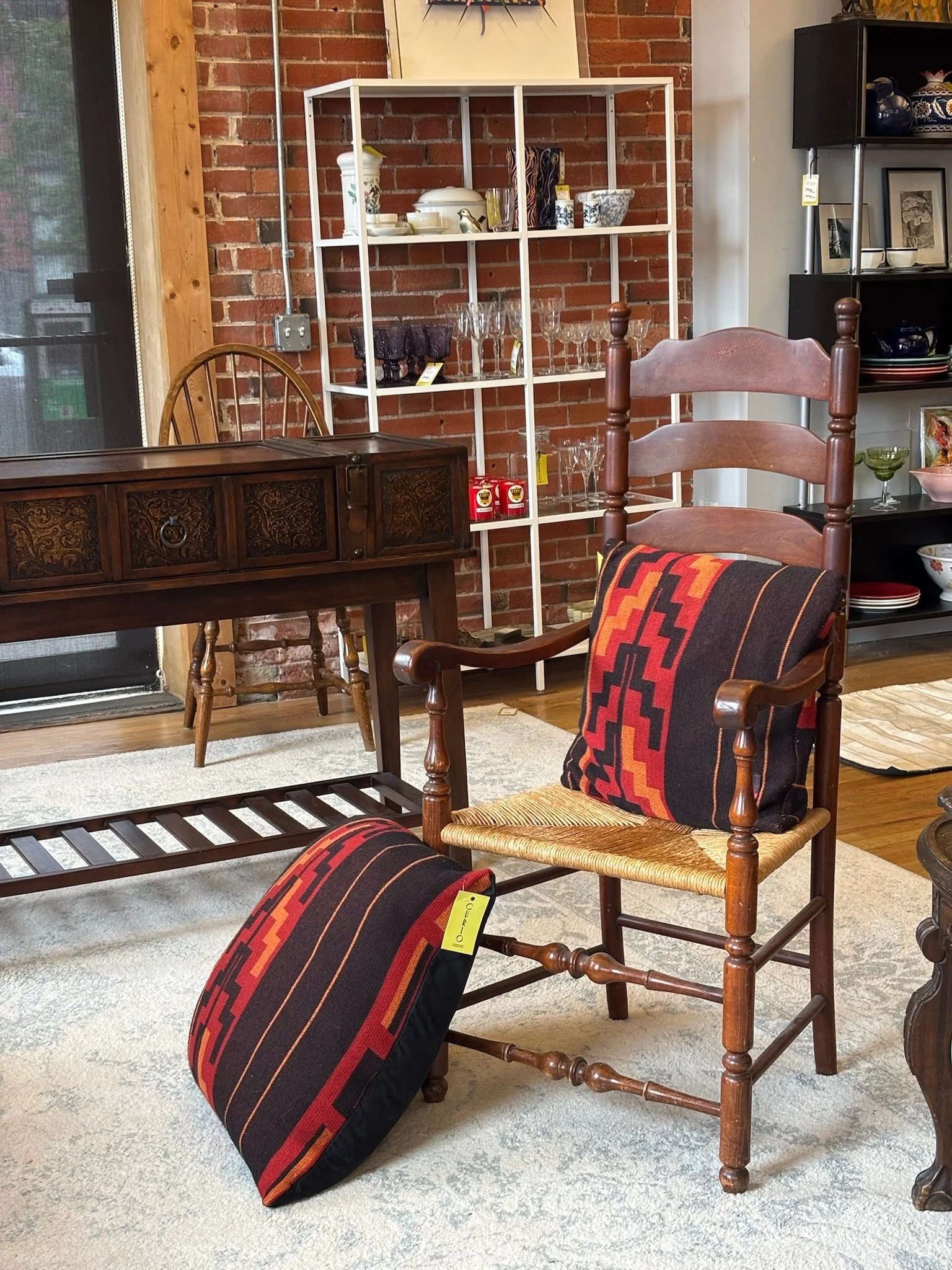 Living room with wooden furniture, a white metal shelf filled with dishes and glassware, a dark wooden cabinet, a black shelving unit with bowls and framed pictures, a wooden chair with a patterned pillow, and a rug on a hardwood floor.