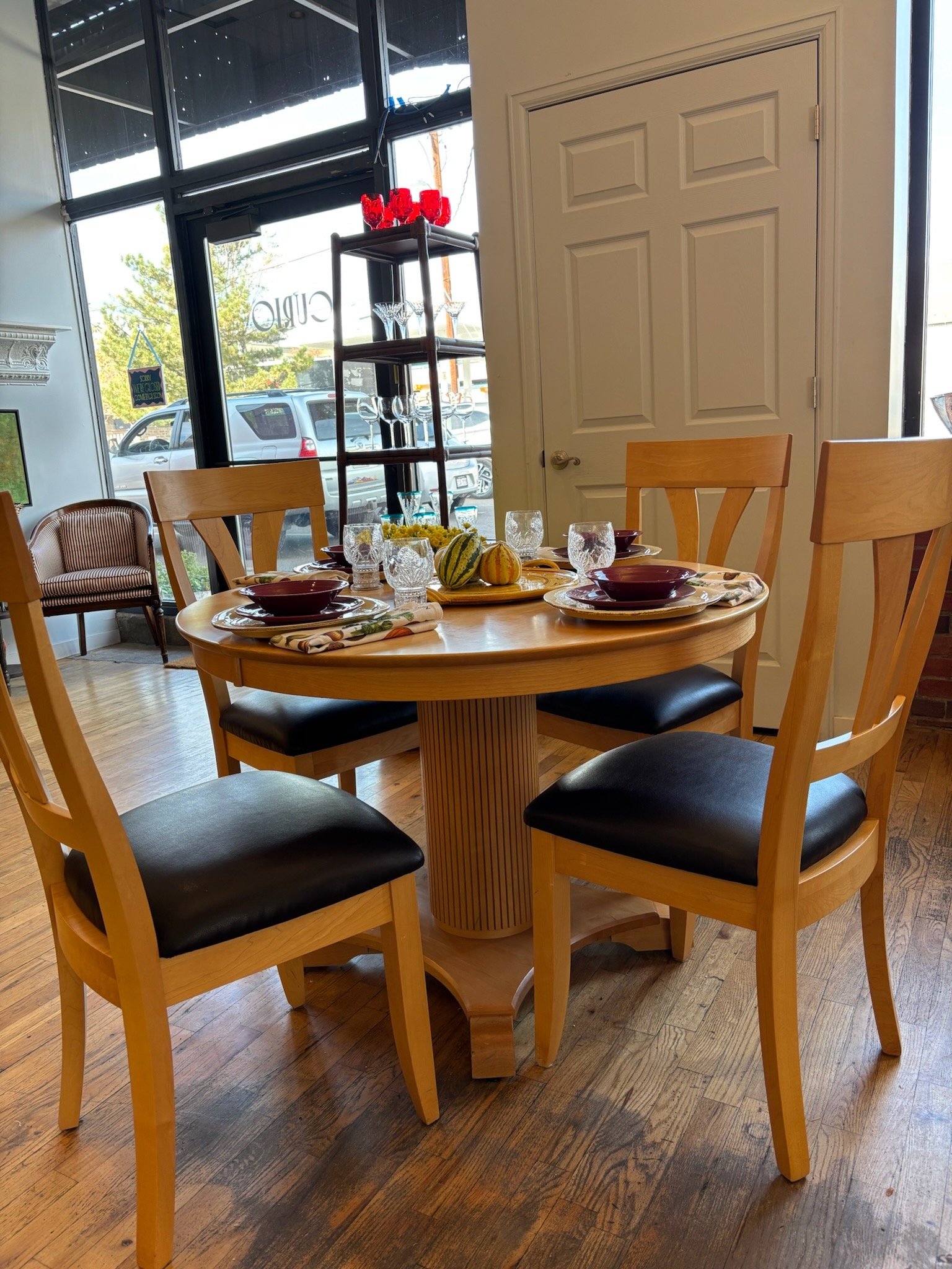 A wooden dining table set with four black-cushioned chairs, four crystal glasses, bowls, and a small tray with decorative gourds, situated in a restaurant with large windows, a white door, and a wooden floor.