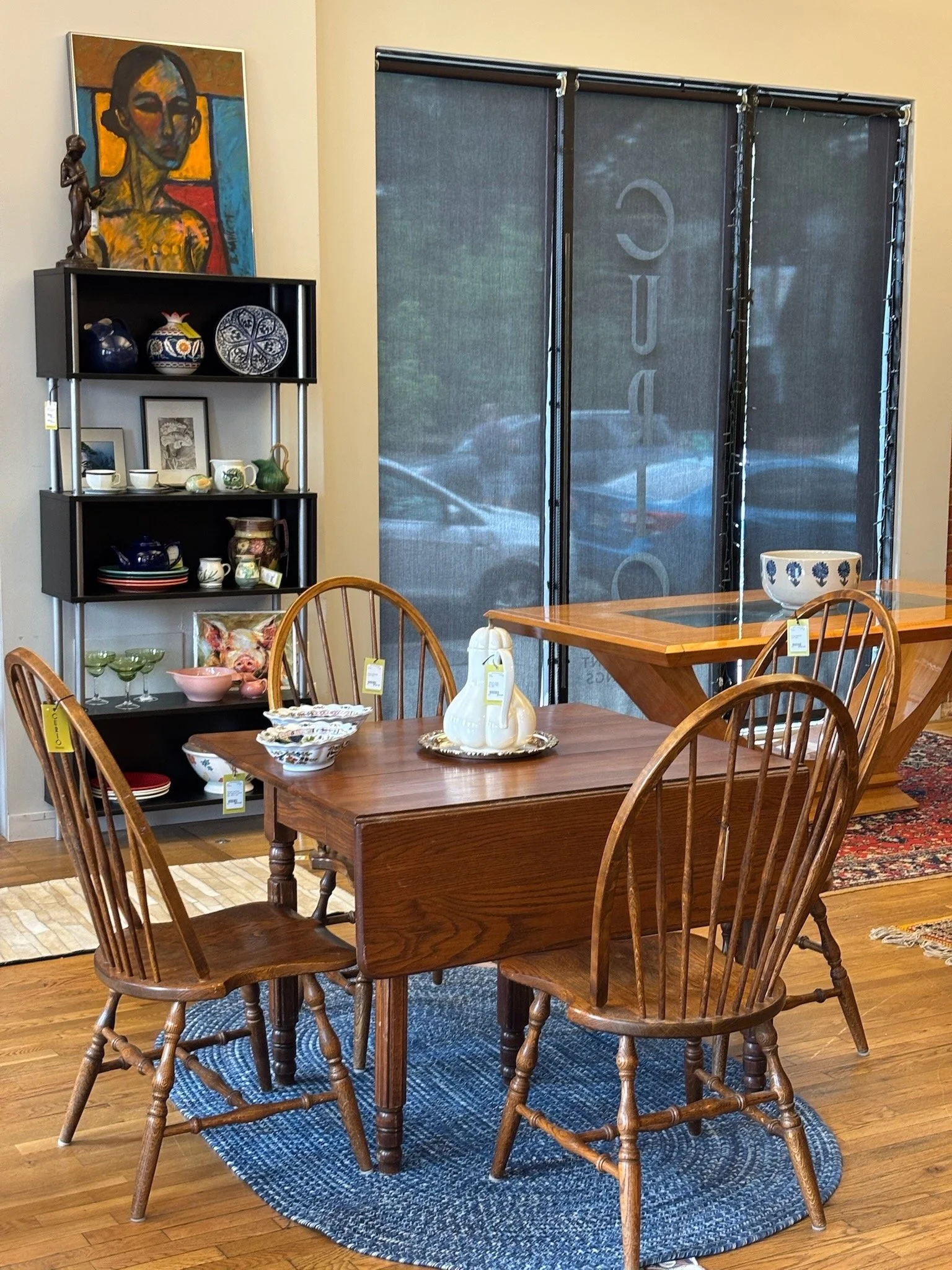A wooden dining table with six matching wooden chairs on a blue round rug. The table has a white pumpkin-shaped decoration and porcelain plates and bowls. In the background, a black shelving unit with decorative ceramics and framed pictures. A colorf