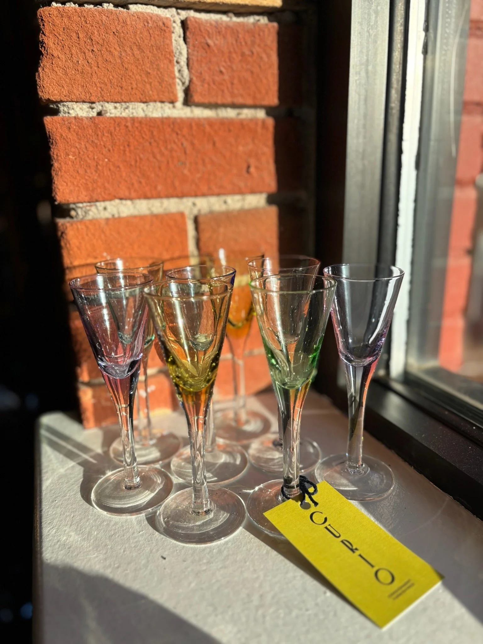 Multiple empty colored champagne glasses placed on a white surface with a brick wall in the background and a yellow tag with black string attached to one glass.