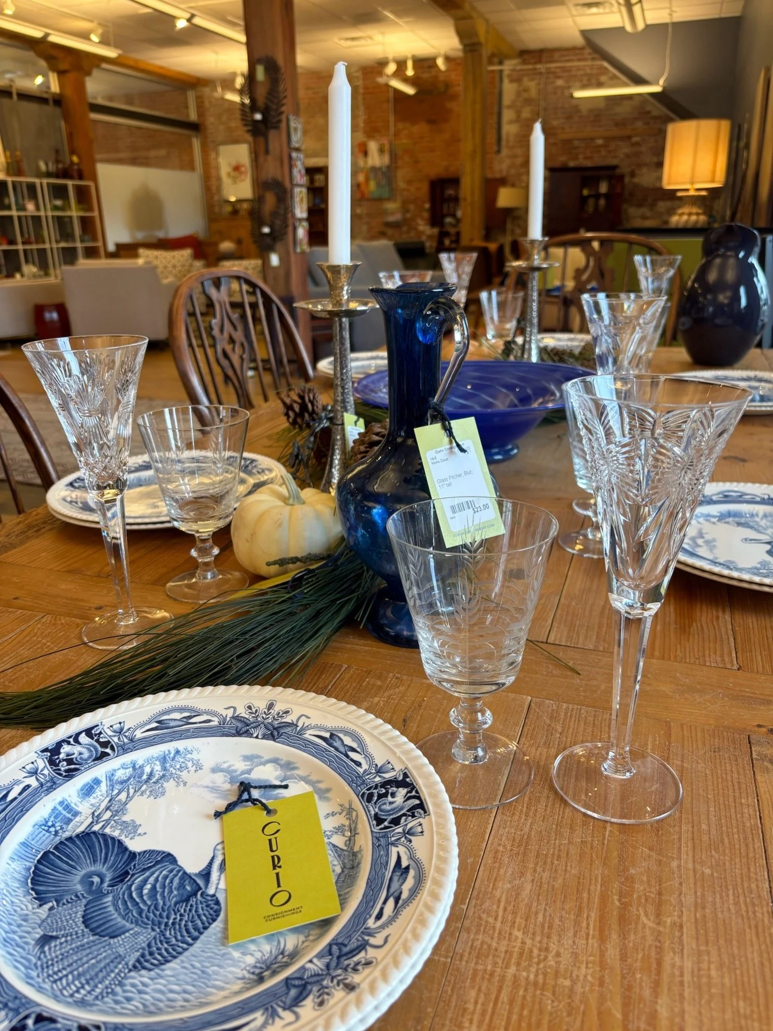 Table set for dining with blue and clear glassware, a blue patterned plate, white pumpkin, pinecones, greenery, and tall white candles in silver candlesticks in a rustic interior