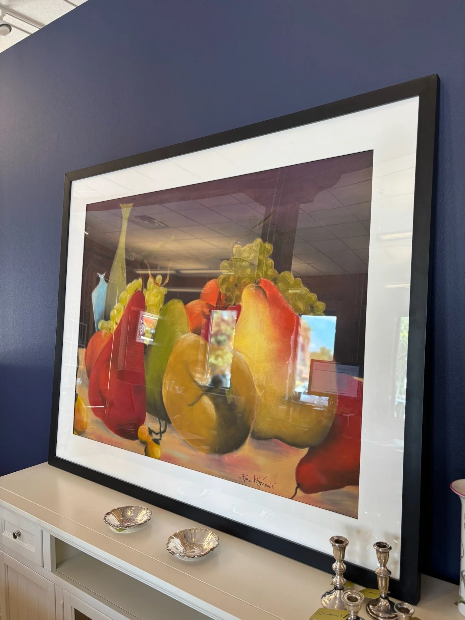 A framed colorful still life painting of various fruits, including grapes, apples, and pears, hangs on a dark blue wall above a white sideboard with three decorative bowls and three silver candle holders.