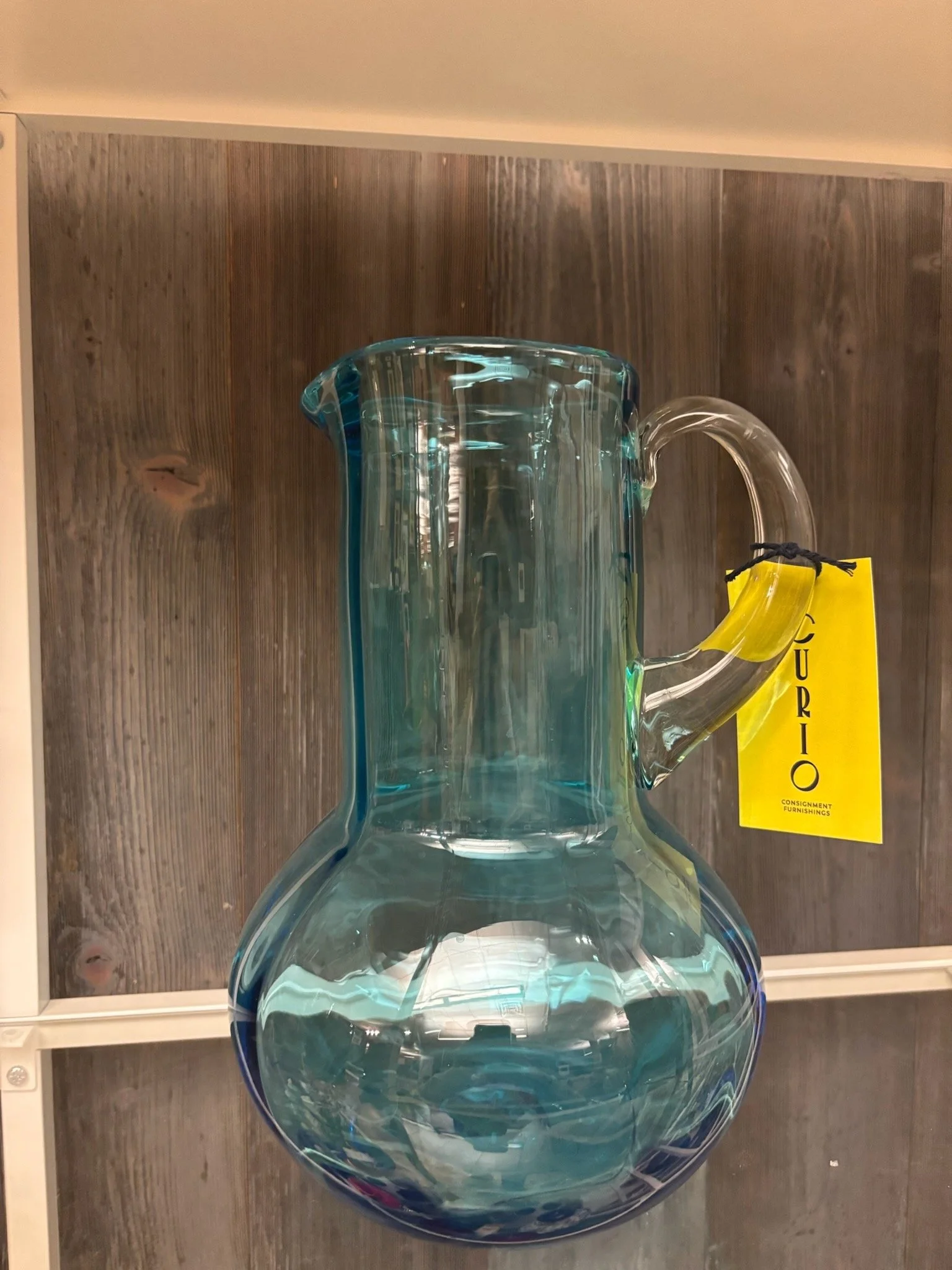 Clear glass pitcher on a wooden shelf with a yellow tag labeled "FURNISHINGS".