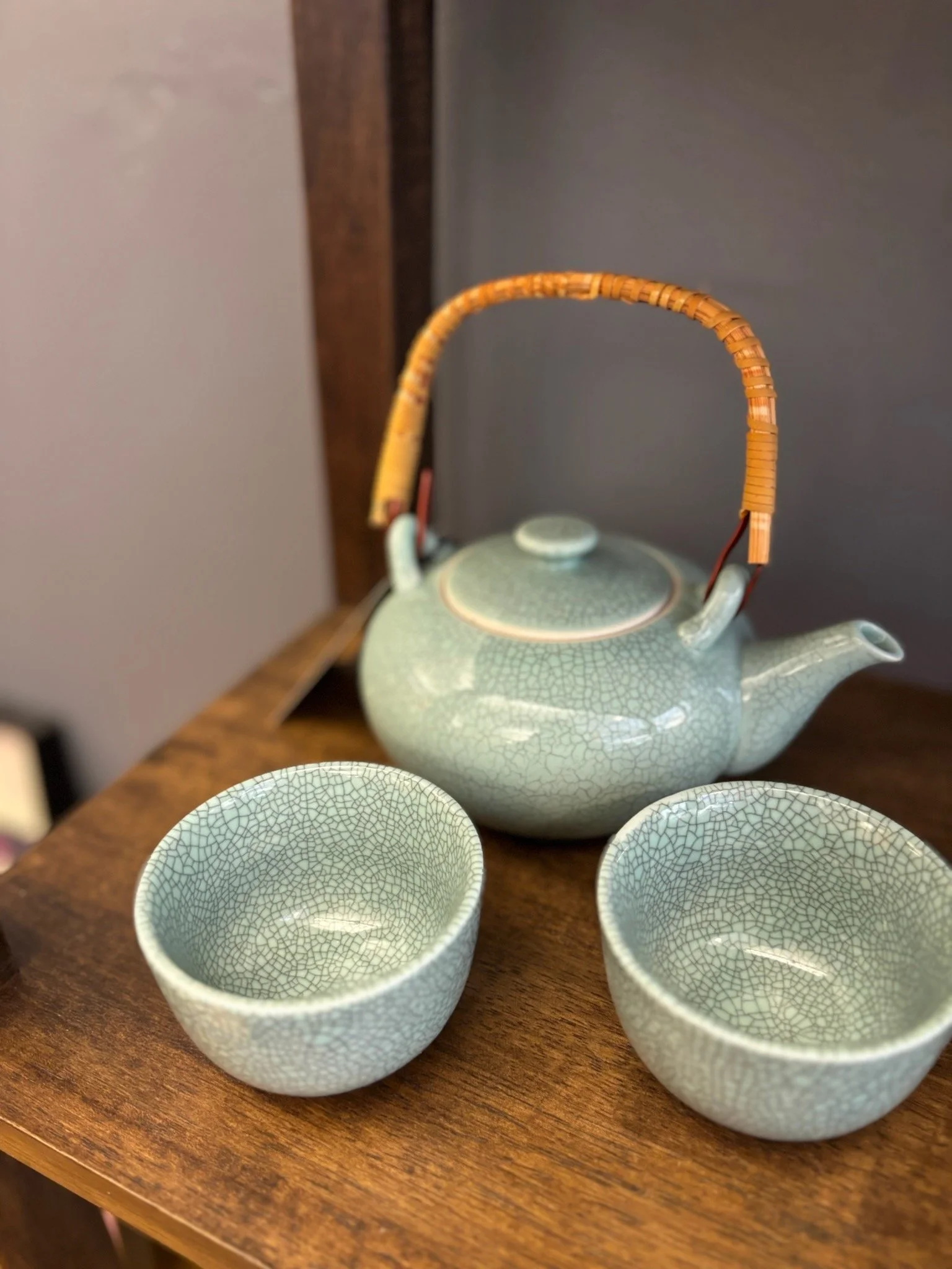 A ceramic teapot with a textured crackled glaze, a woven bamboo handle, and four matching teacups with similar crackled glaze are arranged on a wooden surface.