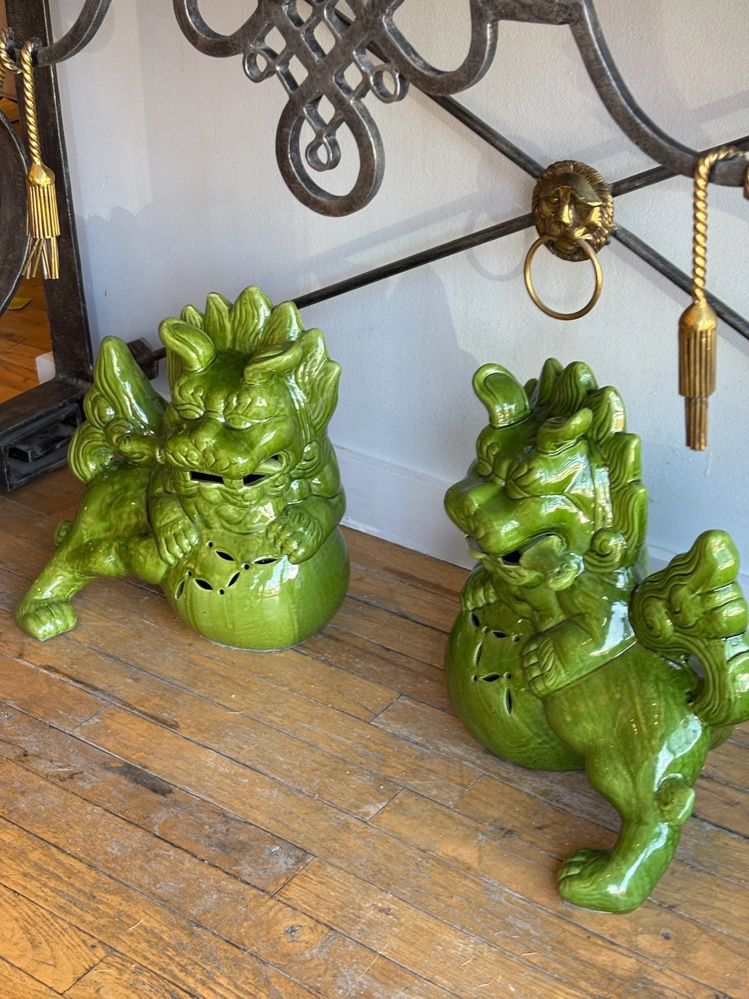 Two green ceramic lion head planters with open mouths, placed on a wooden floor beneath a metal decorative rack with lion head hooks, and a wall in the background.