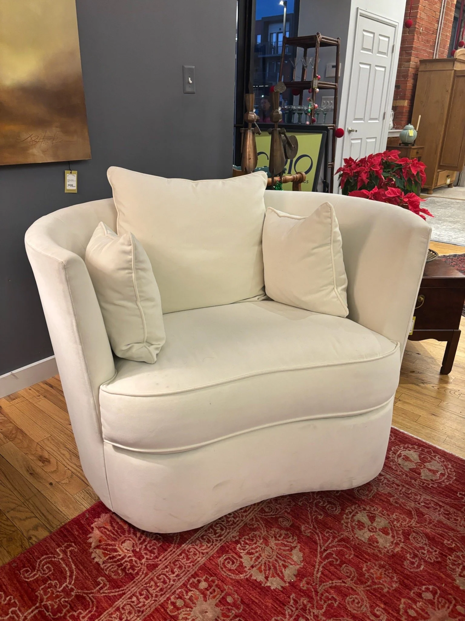 A white upholstered armchair with three matching pillows on a red patterned rug in an interior living space. In the background, there's an art piece on a dark gray wall, a wooden cabinet, and a poinsettia plant next to a white door.