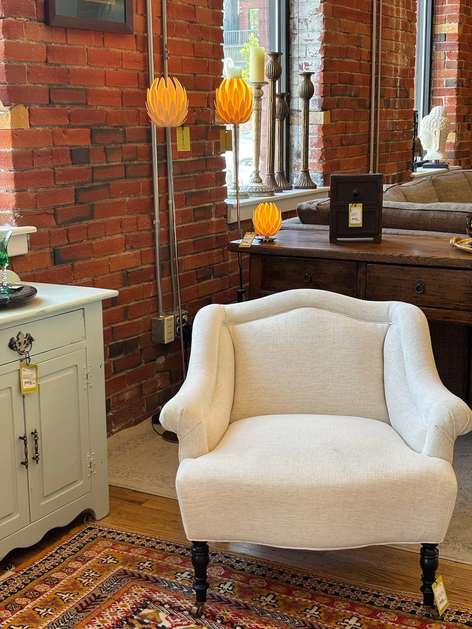 A vintage-style beige armchair with dark wooden legs in a living room with exposed brick walls. A colorful Persian rug is on the floor, and a white cabinet with decorative items is nearby. Behind the chair, a wooden table holds a small lamp and a woo