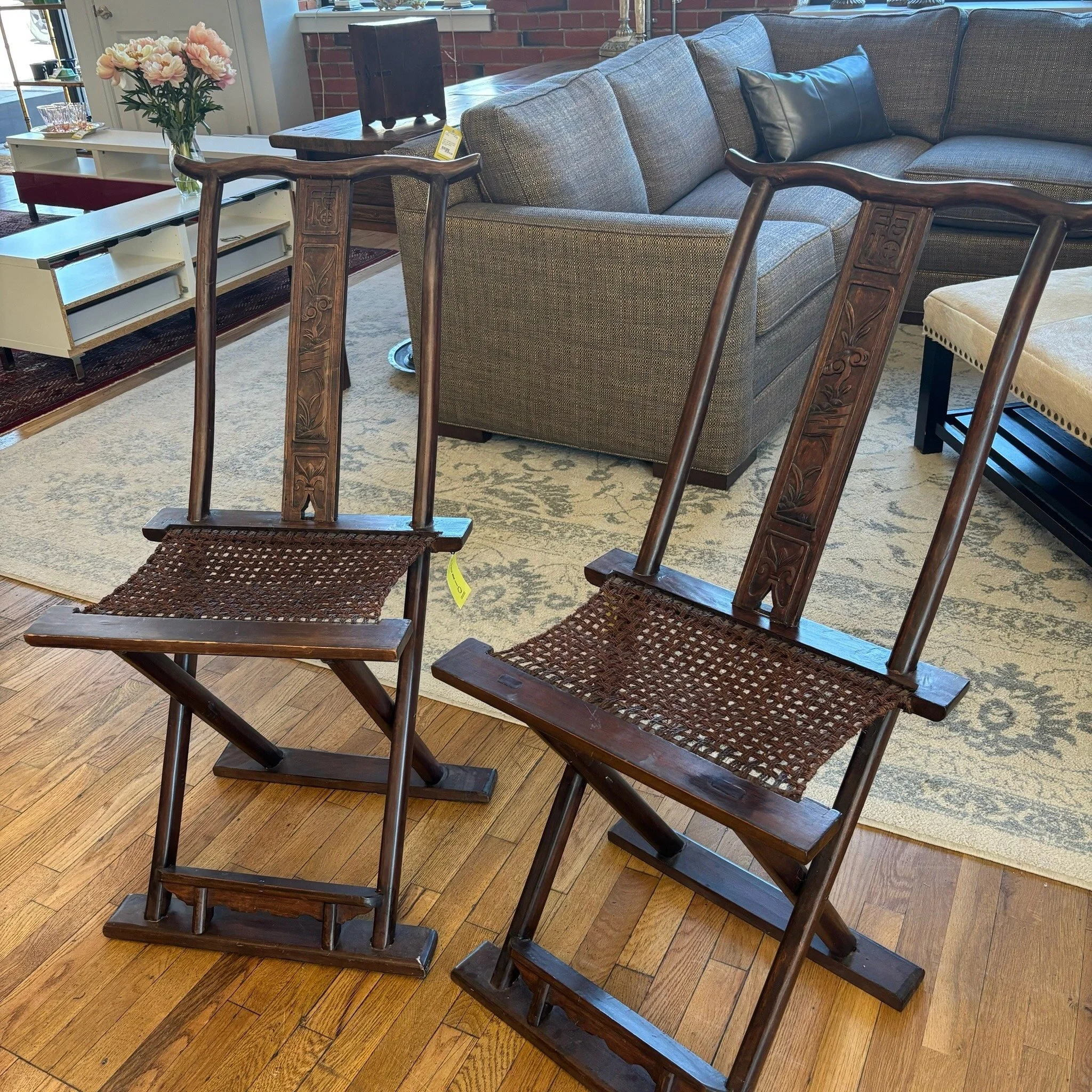 Two wooden chairs with carved designs on the backrests, positioned in front of a living room sofa with cushions, in a space with hardwood flooring and a patterned rug.