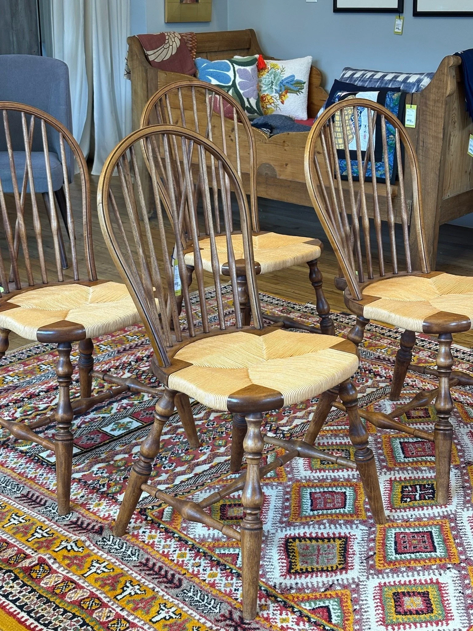 A display of wooden furniture on a colorful patterned rug in a store. Includes a wooden dining table with four matching chairs, a wooden bench with decorative pillows, and a side cabinet, with framed pictures on the wall.