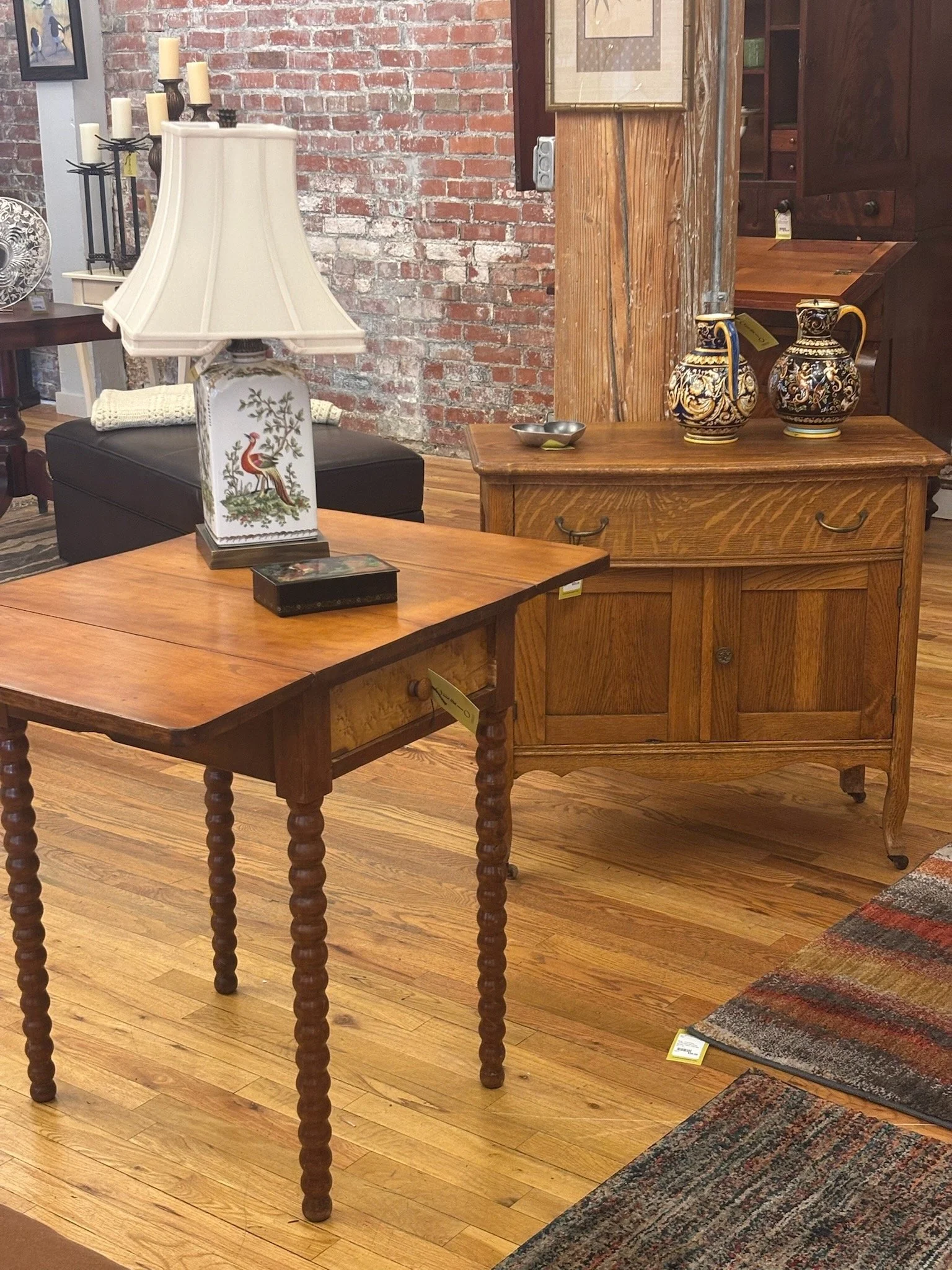 Interior of a furniture store with wooden tables, a wooden cabinet, a table lamp with a cream-colored shade, decorative vases, and a colorful rug on hardwood flooring, exposed brick wall in the background.