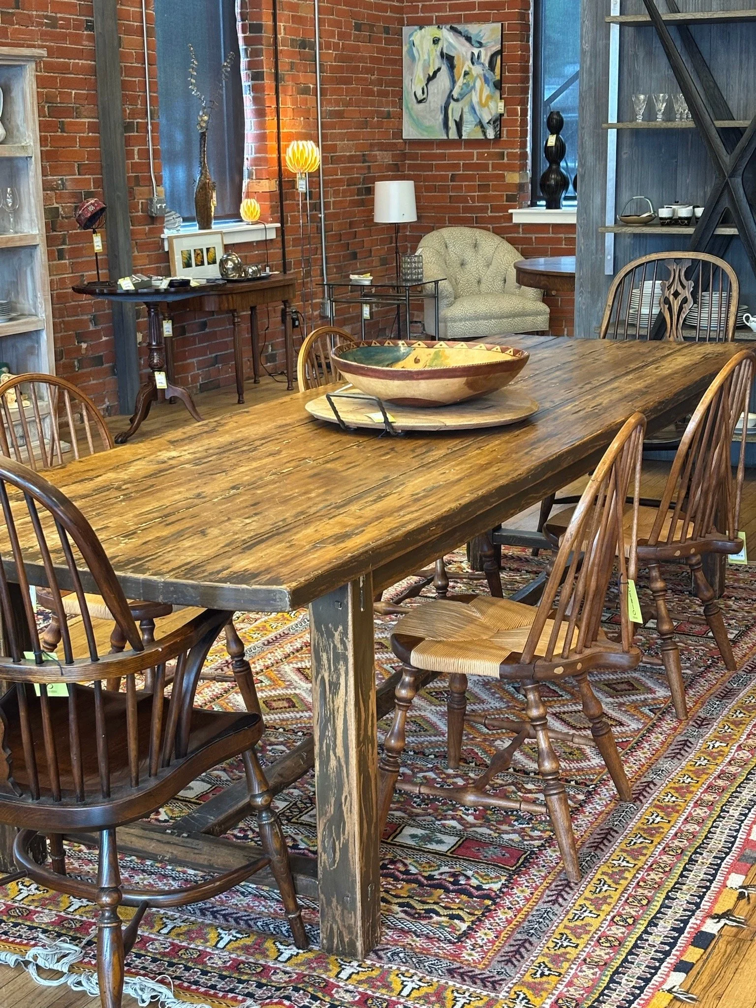A dining room or showroom displaying furniture and decor, including a large wooden dining table with chairs, a patterned rug, a corner armchair, side tables, lamps, artwork, and decorative objects against exposed brick walls.