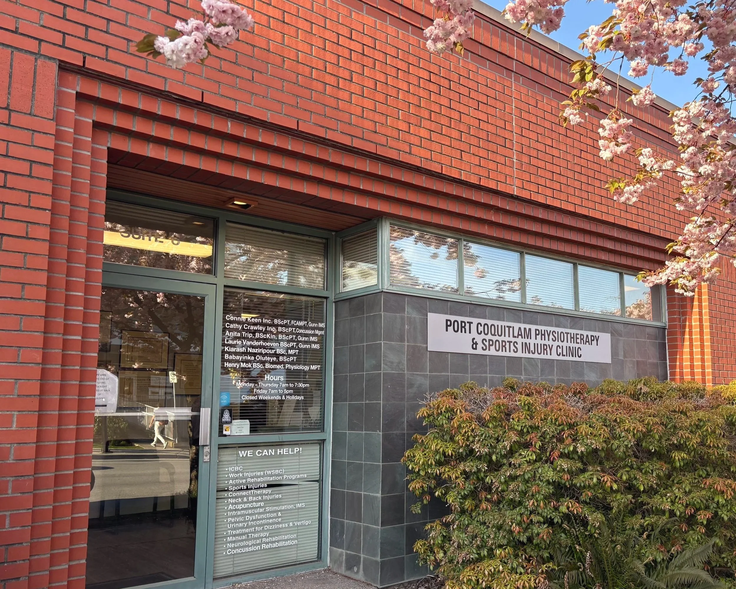 Exterior view of Port Coquitlam Physiotherapy & Sports Injury Clinic, with a glass door and sign, surrounded by green bushes and brick and tiled walls.