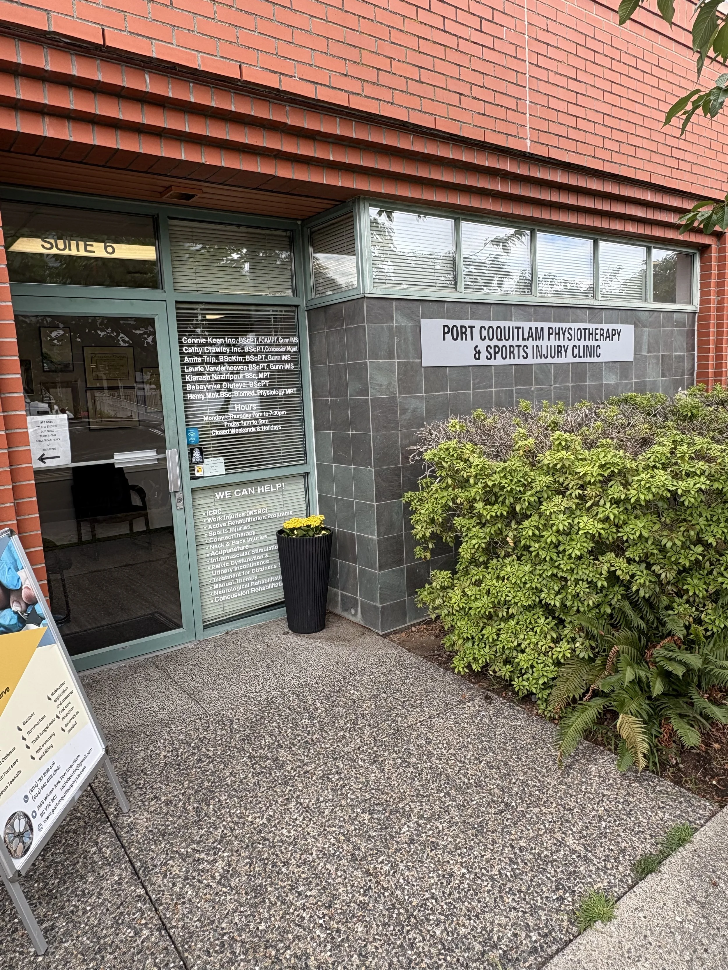 Exterior view of Port Coquitlam Physiotherapy & Sports Injury Clinic, with a glass door and sign, surrounded by green bushes and brick and tiled walls.