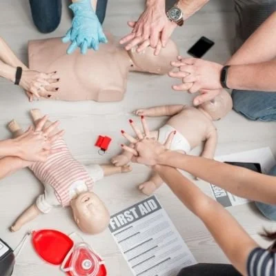 Group practicing CPR on adult and infant mannequins, hands shown applying chest compressions, with first aid materials nearby.