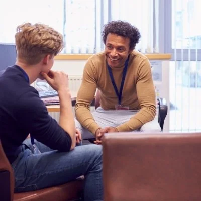 A man sitting in a chair facing another person, possibly in an office setting, engaged in a conversation.