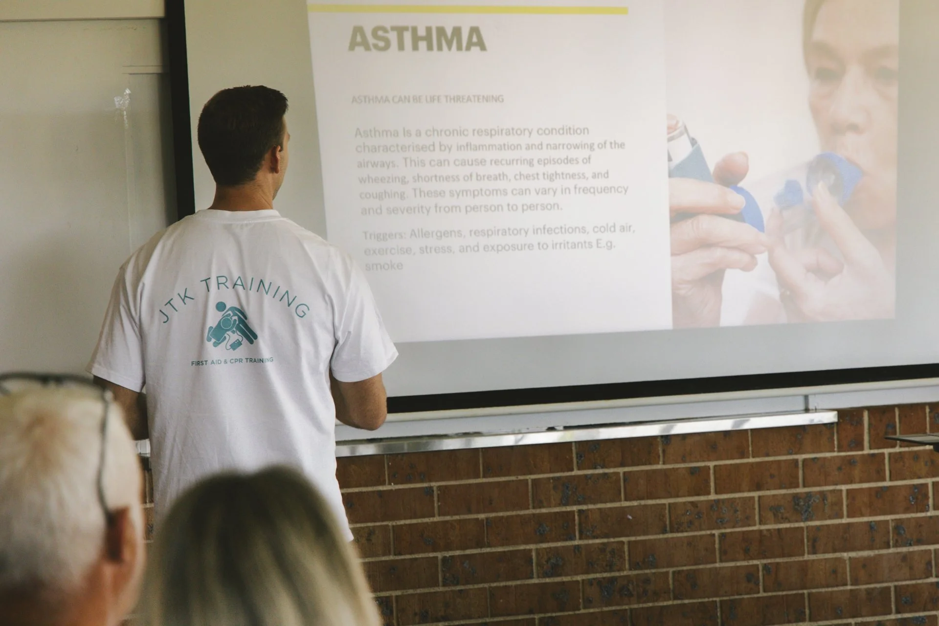 A person in a JTK Training shirt standing in front of a screen displaying a presentation slide about asthma. The slide contains information about asthma triggers and symptoms, and includes an image of a person using an inhaler. There are several people seated, facing the presentation.