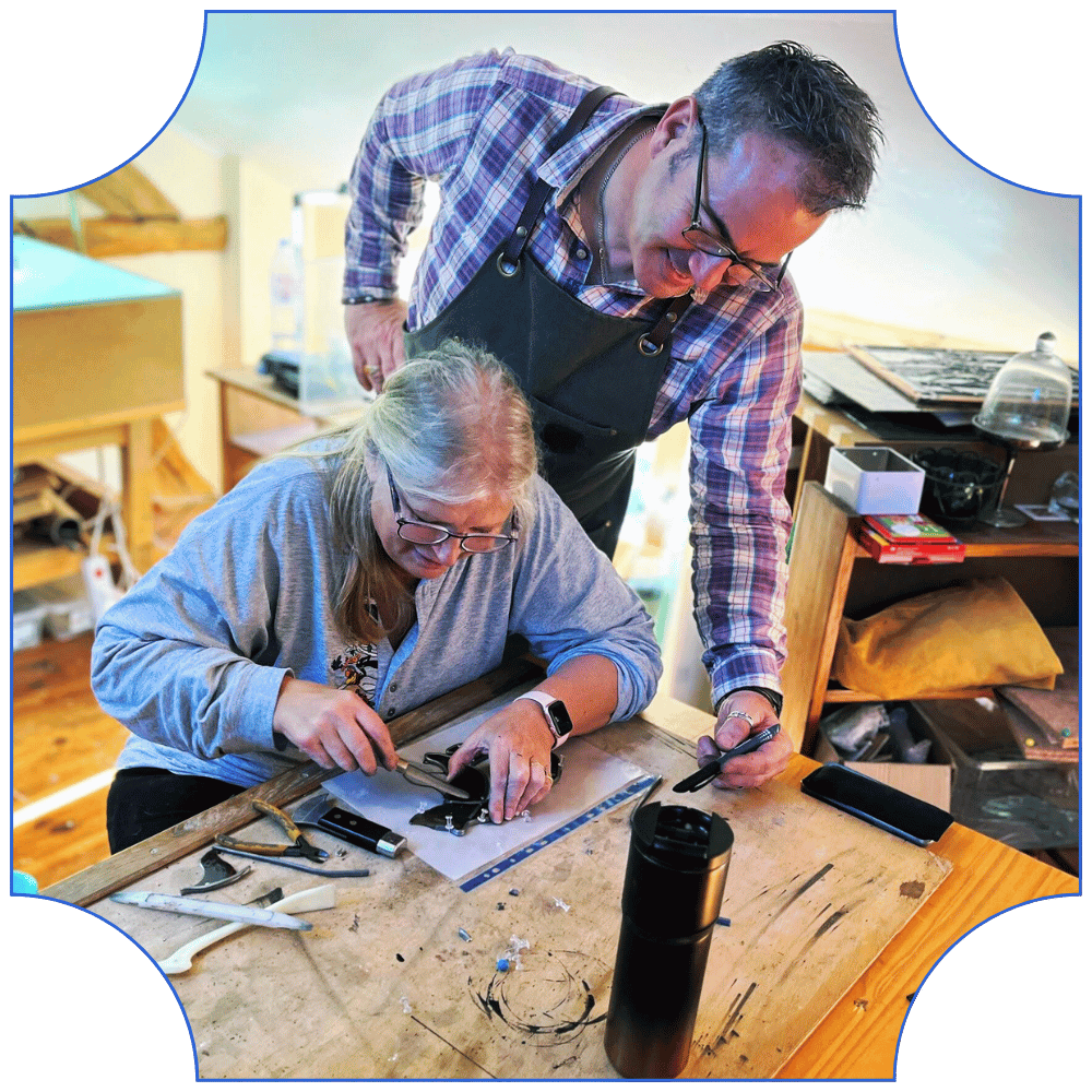 David Tweddle, artist at Lumiere Stained Glass Art and Training Studio, guiding a student as she solders a stained glass piece at the workbench, surrounded by tools and materials in the studio.