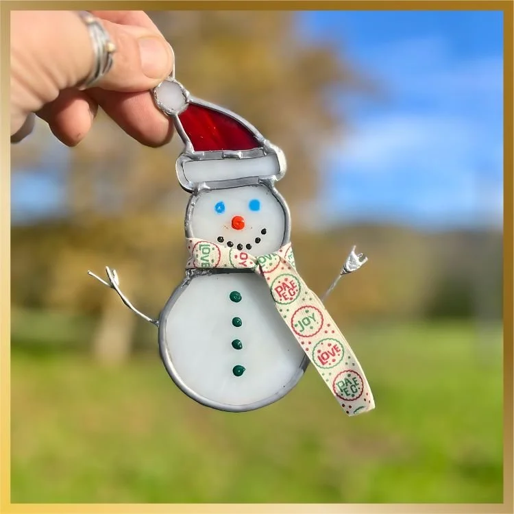 Hand holding a small stained glass snowman ornament wearing a striped scarf and Santa hat, outdoors with a blurred green field background.