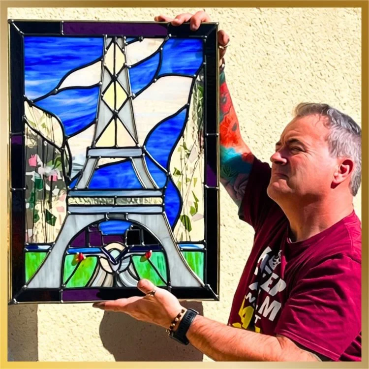 David holding a student’s finished stained glass panel featuring the Eiffel Tower against a blue sky, with green fields below | Lumiere Stained Glass Art & Training