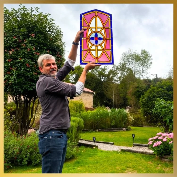 Student Andy holding a finished stained glass window panel with a pink and blue geometric medallion design, outdoors in a garden | Lumiere Stained Glass Art & Training