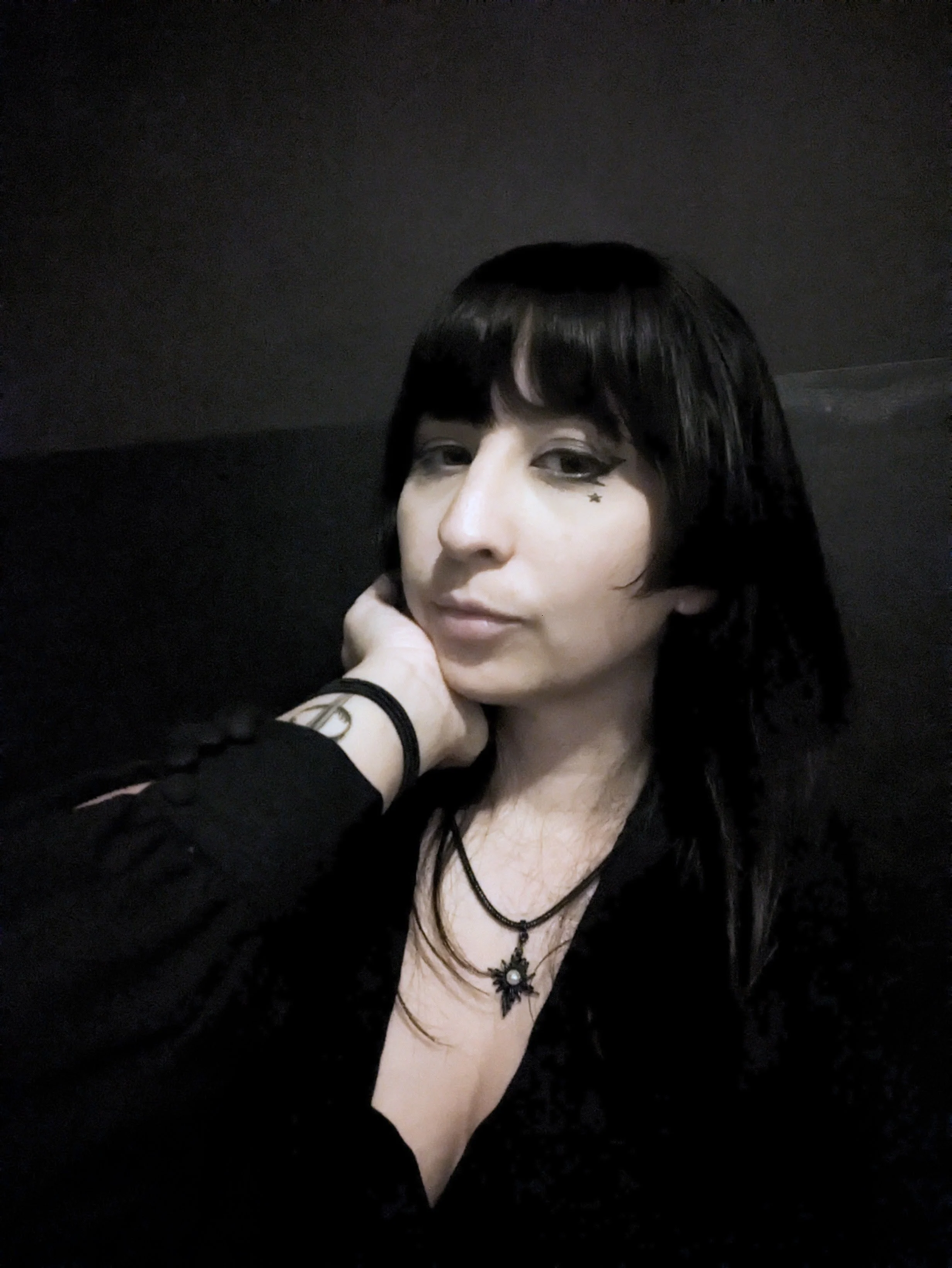 A person with dark hair and makeup, wearing black clothing, multiple necklaces including one with a winged pendant, and a star tattoo near their eye, resting their face on their hand against a dark background.