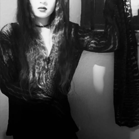 Black-and-white photo of a woman with long wavy hair, wearing a lace top and choker, holding a garment on her arm.