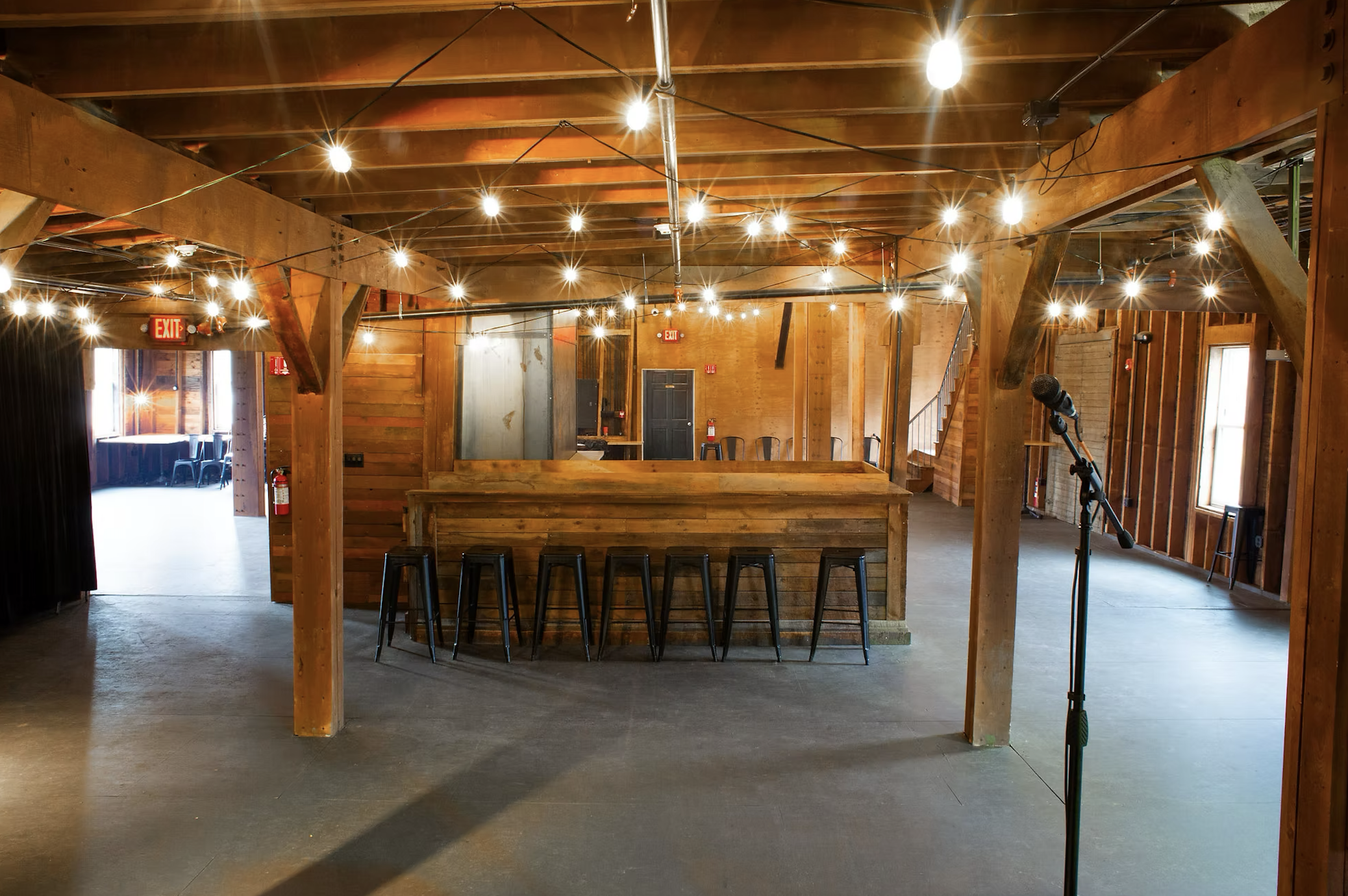 An empty event venue with a wooden bar and string lights hanging from the ceiling, microphone stand, and open doorway with chairs in the background.