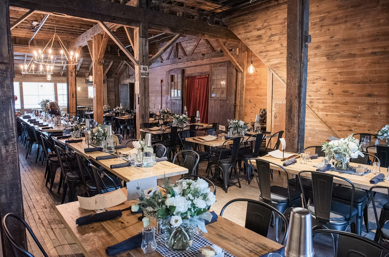 A rustic wooden event space with long tables decorated with floral centerpieces, table settings, and water bottles, set for a celebration or gathering.