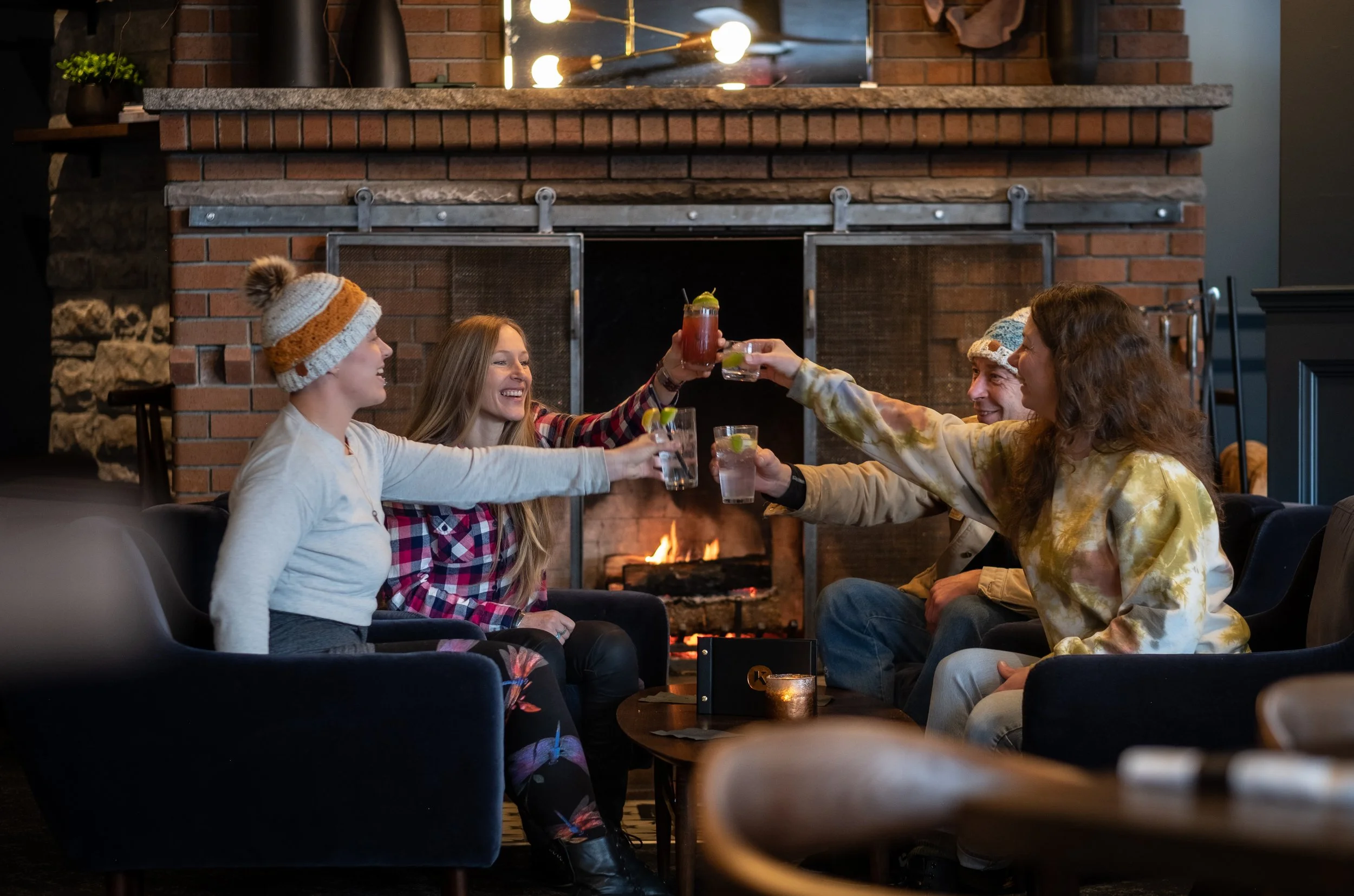Group of four friends sitting on couches by a fireplace, raising drinks for a toast, smiling, in a cozy indoor setting.
