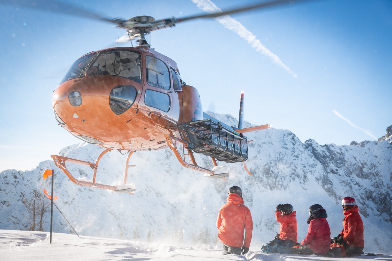 A helicopter flying over a snowy mountain landscape with four people dressed in red gear sitting on the snow, observing the scene.