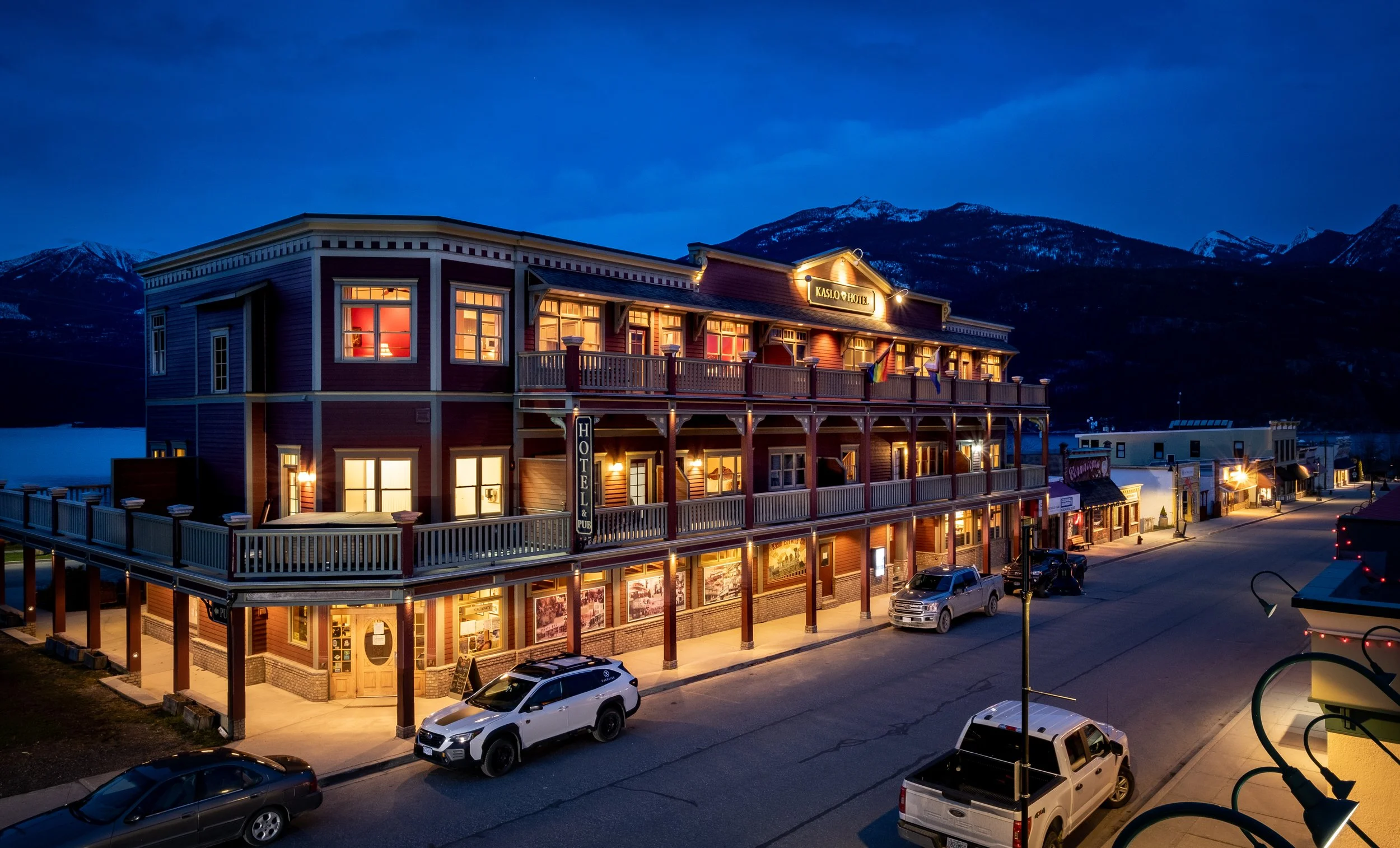 Nighttime view of a three-story hotel with warm interior lights, wooden balconies, and a sign that says 'Kaslo Hotel.' The hotel is on a street with parked cars, and mountains are visible in the background under a dark twilight sky.