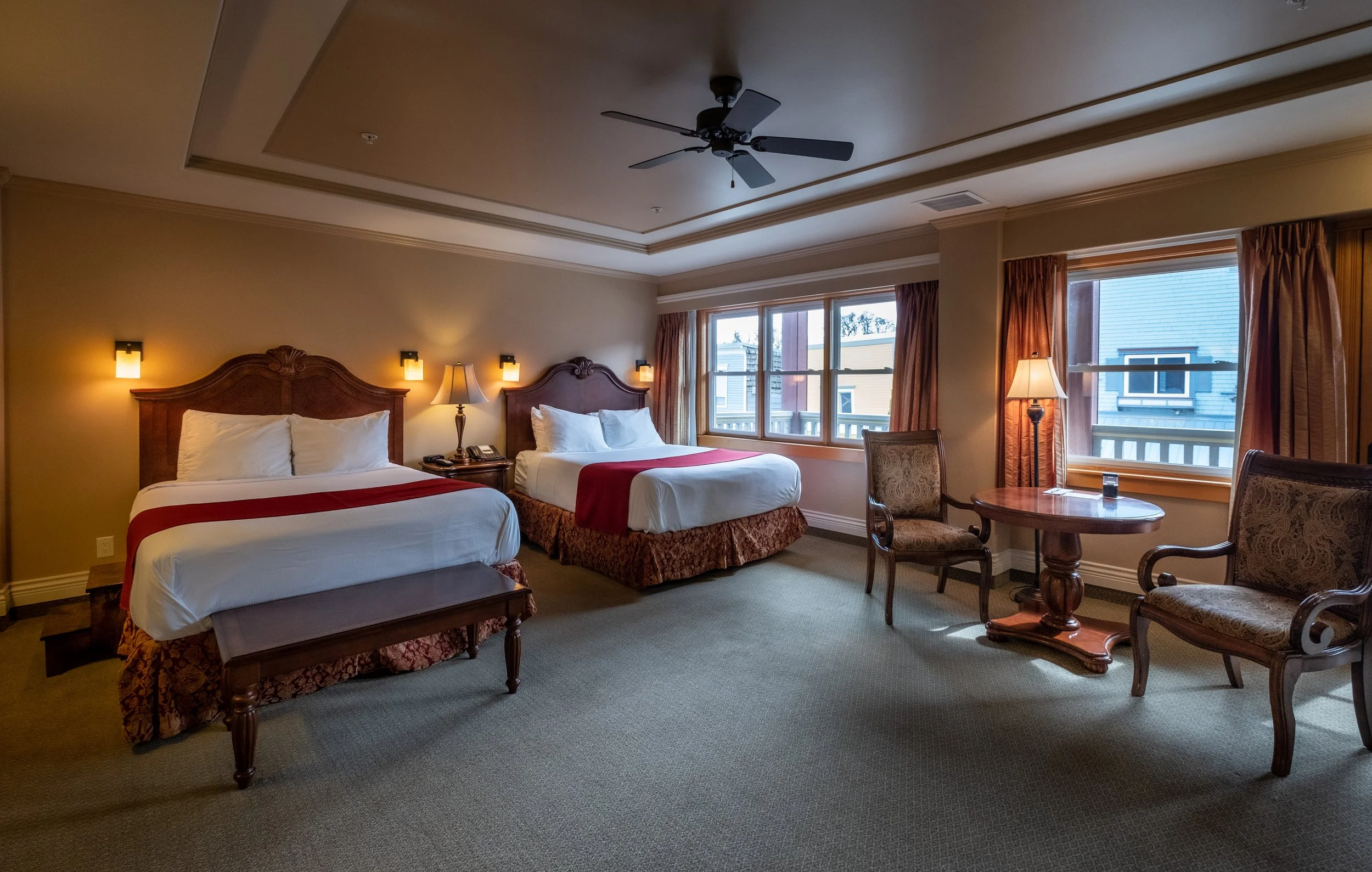 Hotel room with two beds, lamps, chairs, a round table, and large windows showing neighboring houses.
