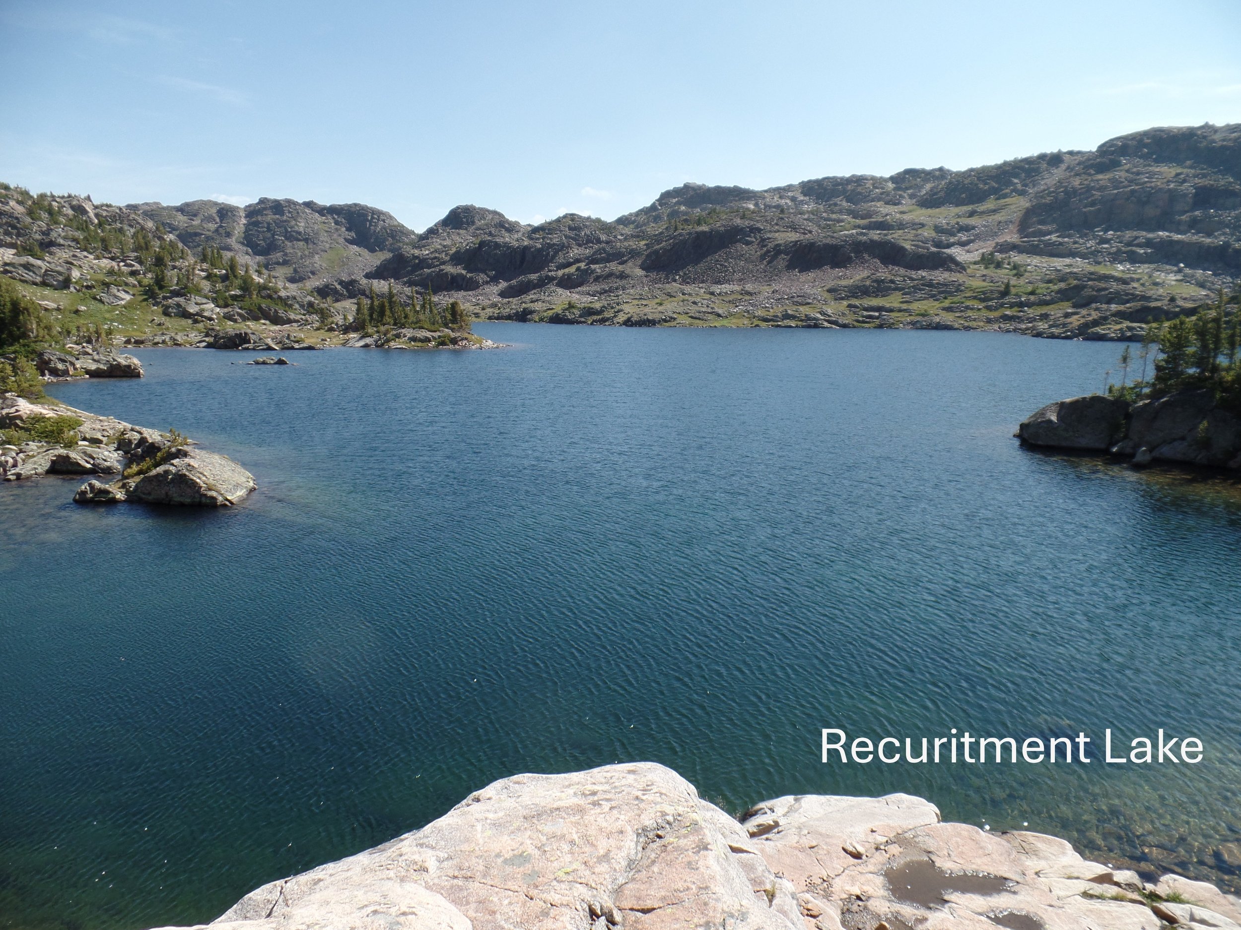 Mountain lake surrounded by rocky hills and evergreen trees under a clear sky, labeled as "Recruitment Lake."