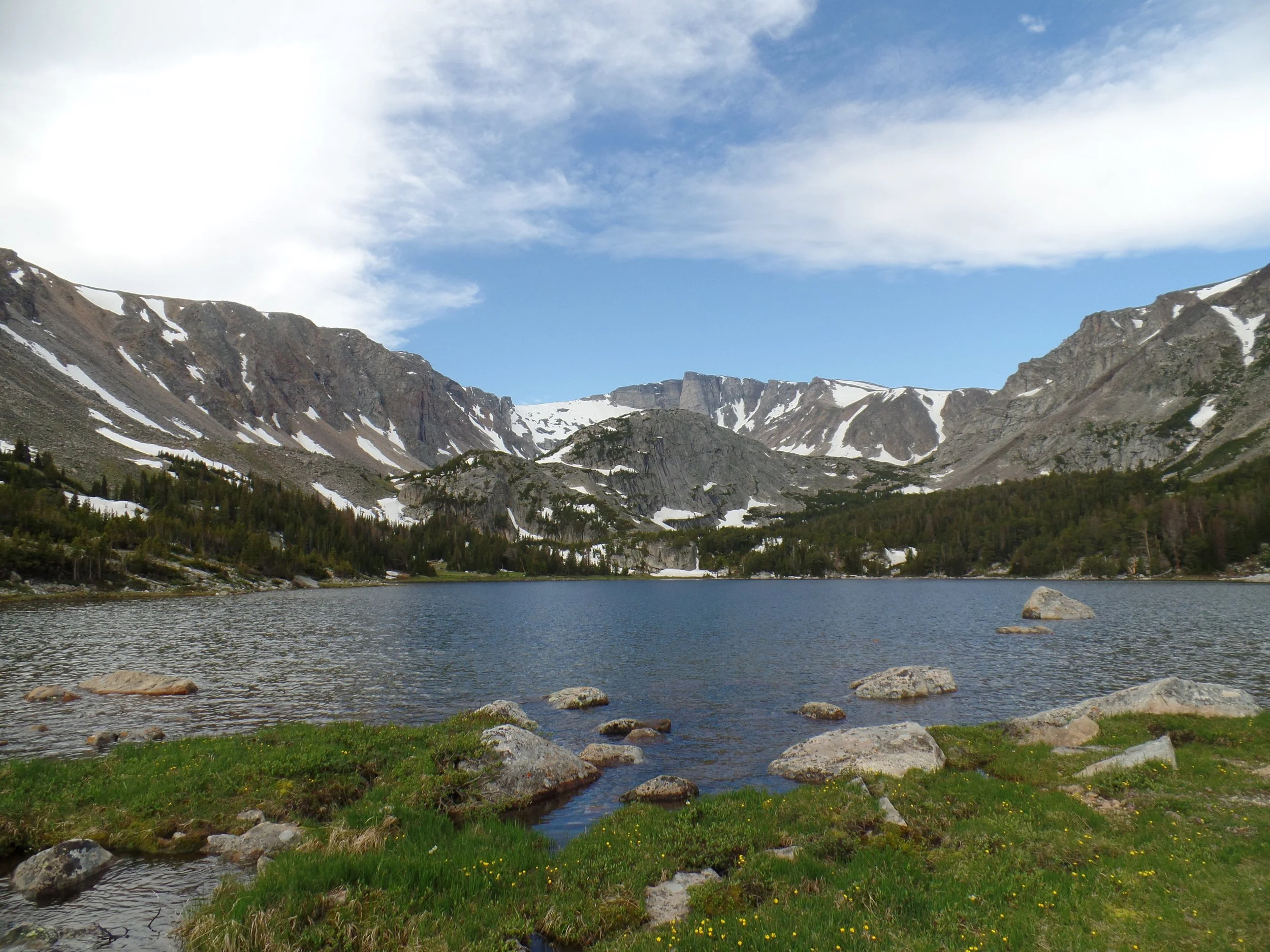 Mountain lake with surrounding snow-capped peaks and green grass, under a partly cloudy sky.