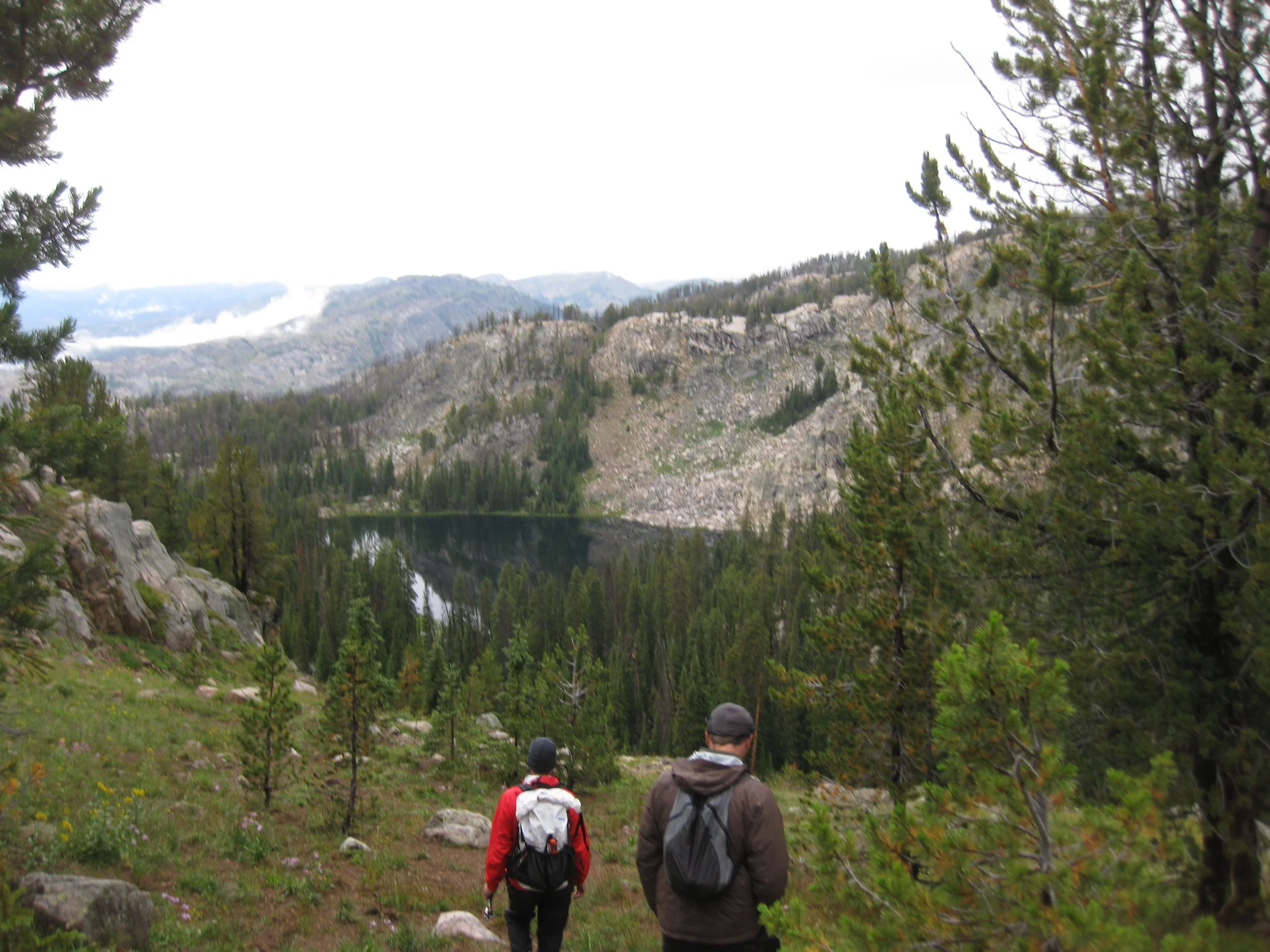 Two hikers descend a rocky slope with a scenic view of a forested valley and distant mountains.