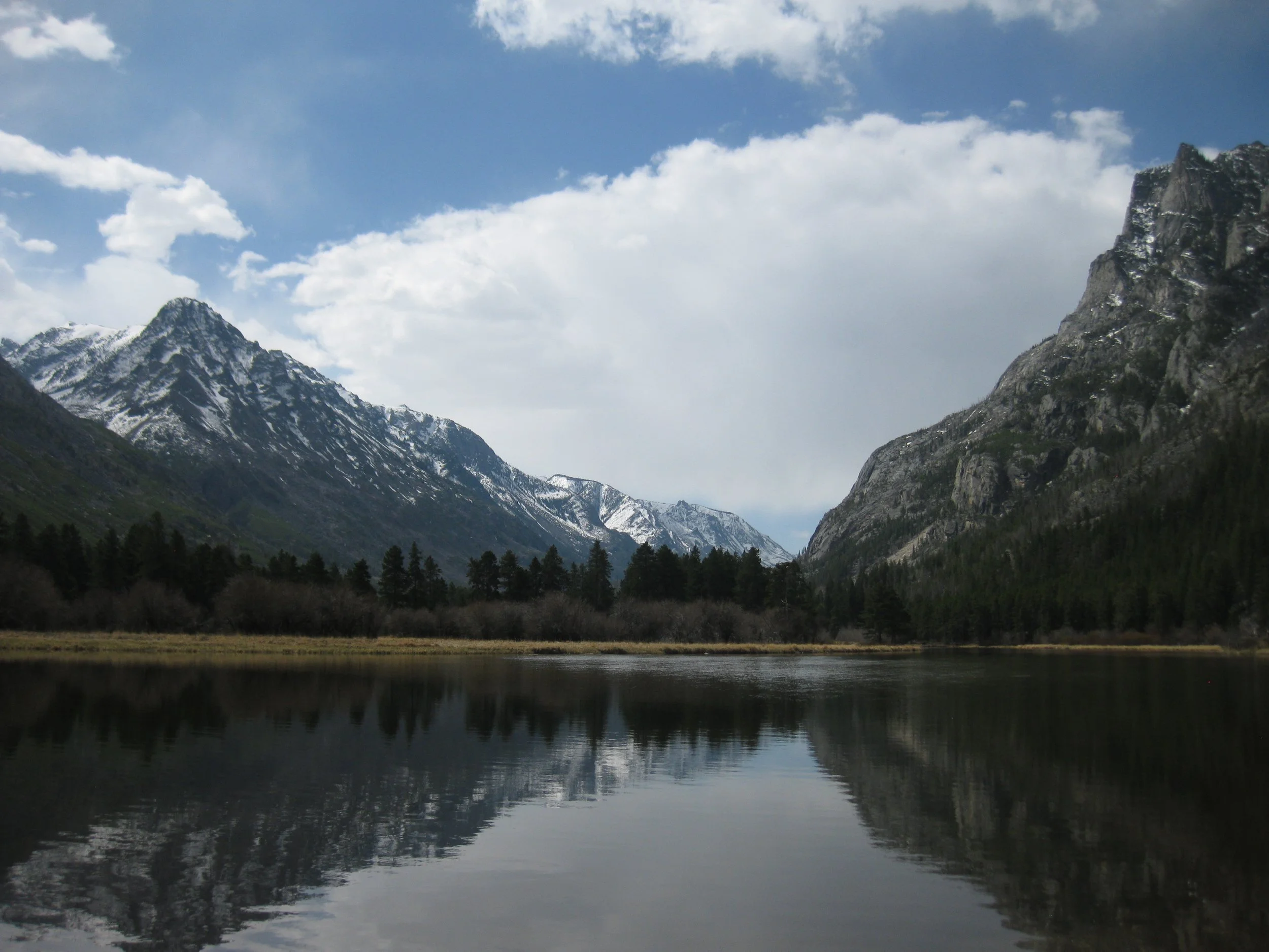 Mountain lake with snow-capped peaks, pine trees, and clouded sky reflection.