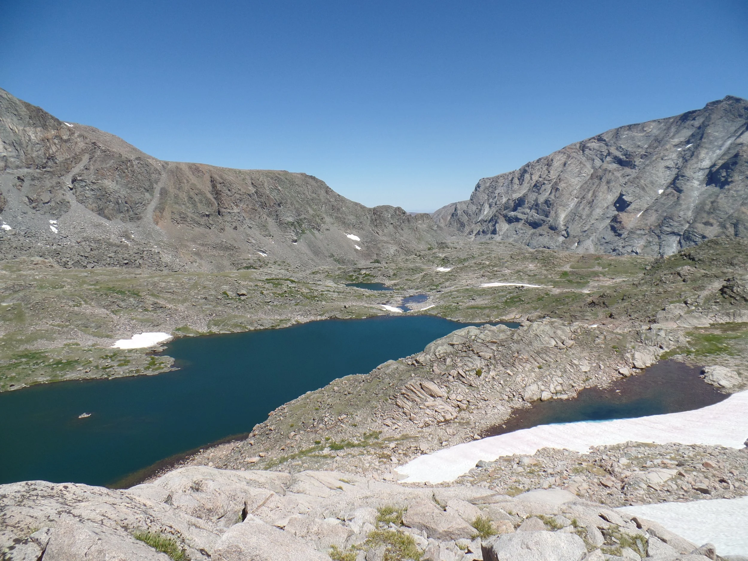 Mountainous landscape with a blue lake, rocky terrain, patches of snow, and clear blue sky.