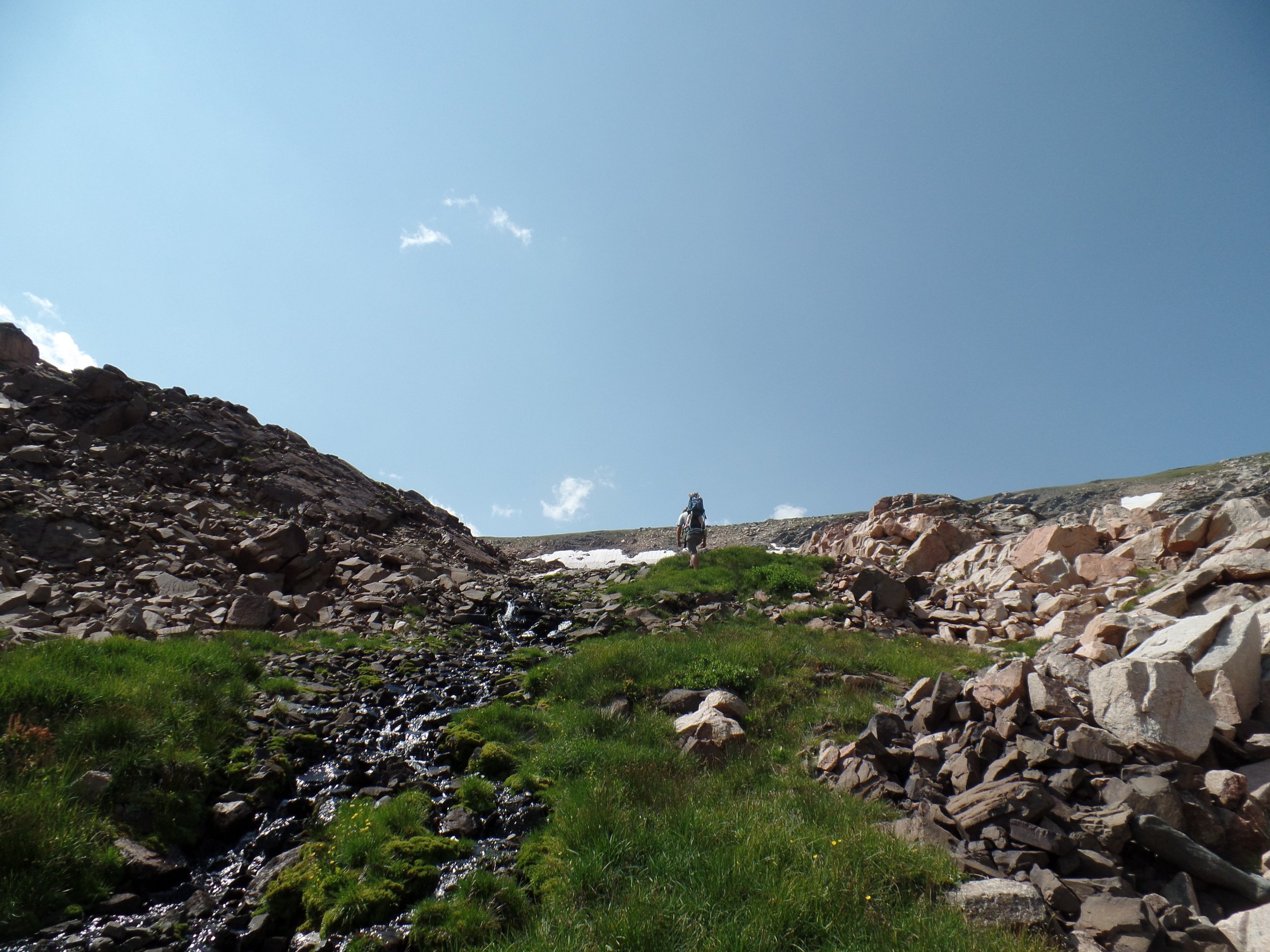 Hiker on rocky terrain with small stream and green grass under blue sky