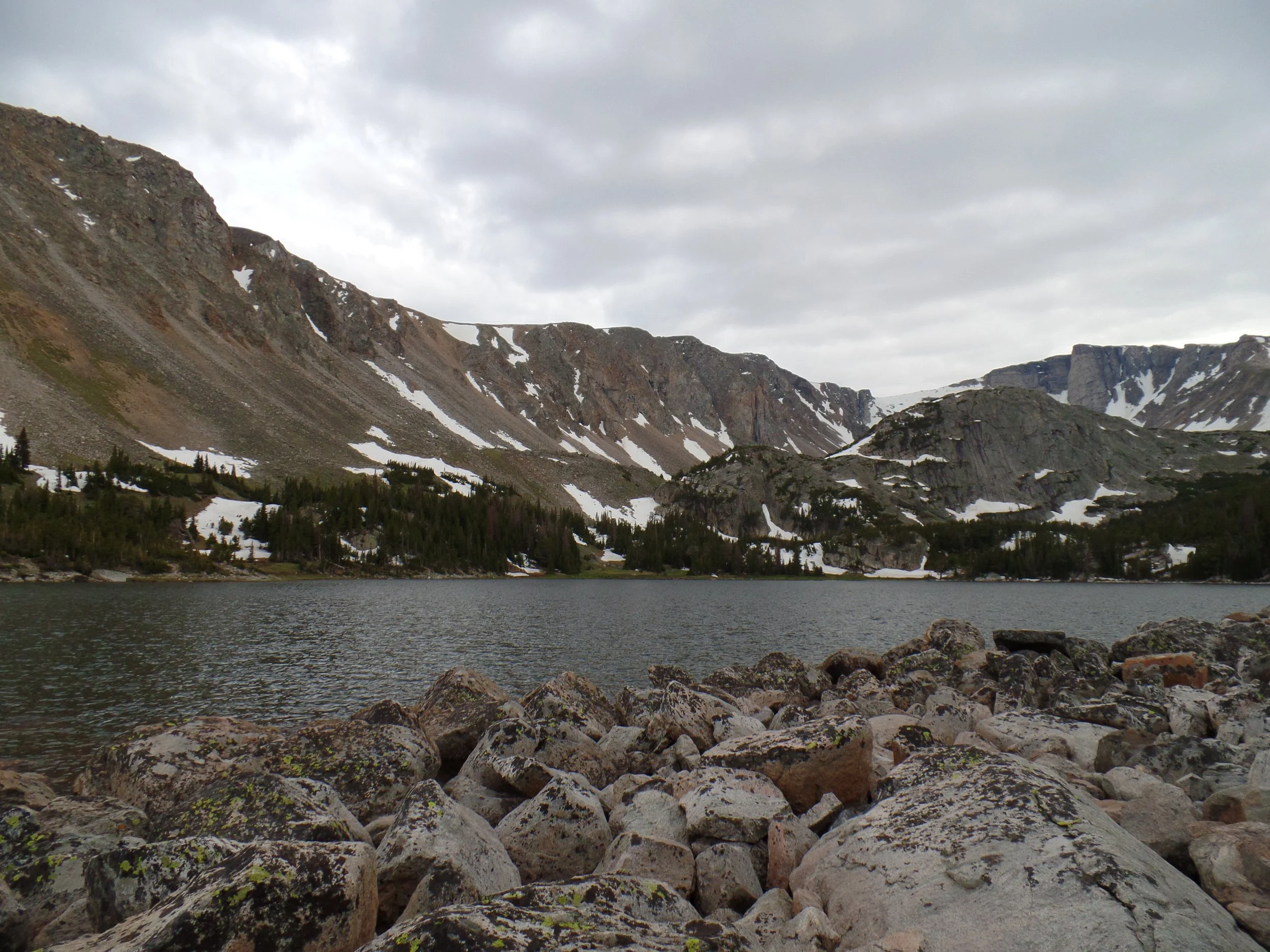 Rocky mountain landscape with snowy patches and a lake, surrounded by rugged cliffs and a cloudy sky.