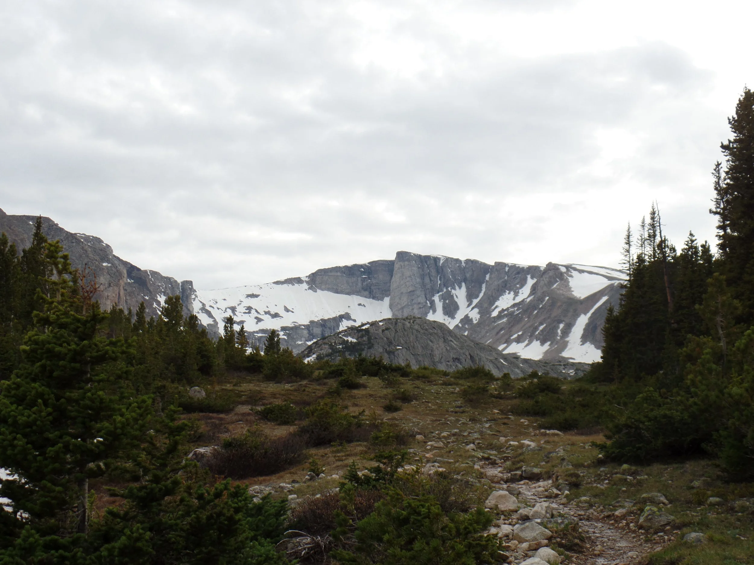 Mountain landscape with snow patches, rocky terrain, and evergreen trees under a cloudy sky.