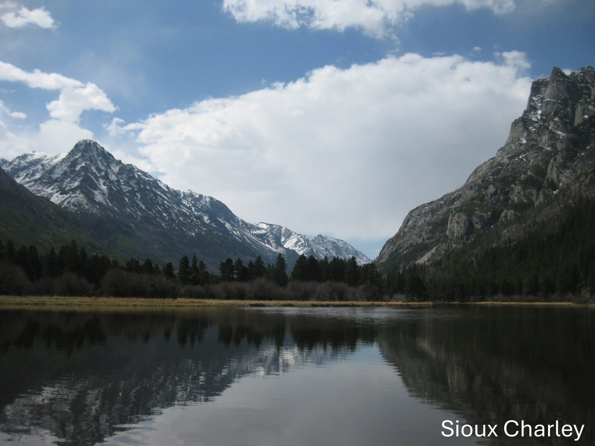 Mountain landscape with a lake reflecting snowy peaks and pine trees, cloudy sky overhead. "Sioux Charley" text at the bottom.