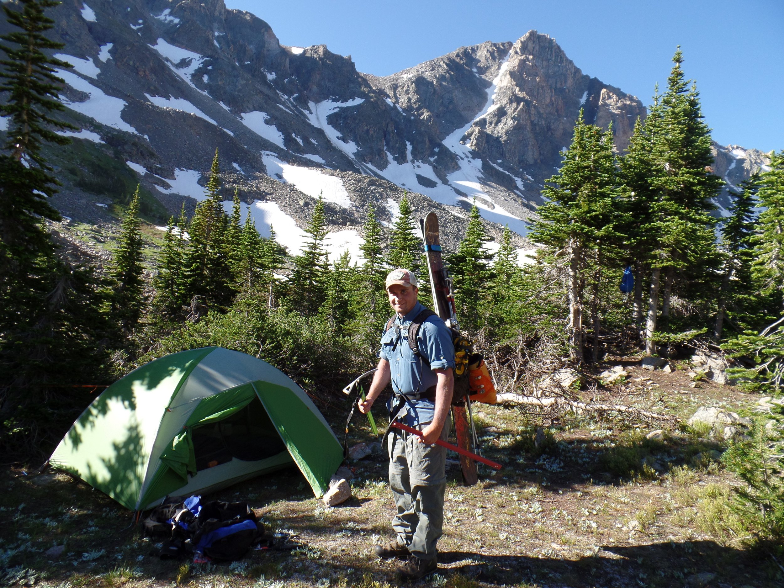 A person standing near a green tent in a mountainous area, holding skiing poles, with skis attached to their backpack. The background features snow-capped peaks and evergreen trees under a clear blue sky.