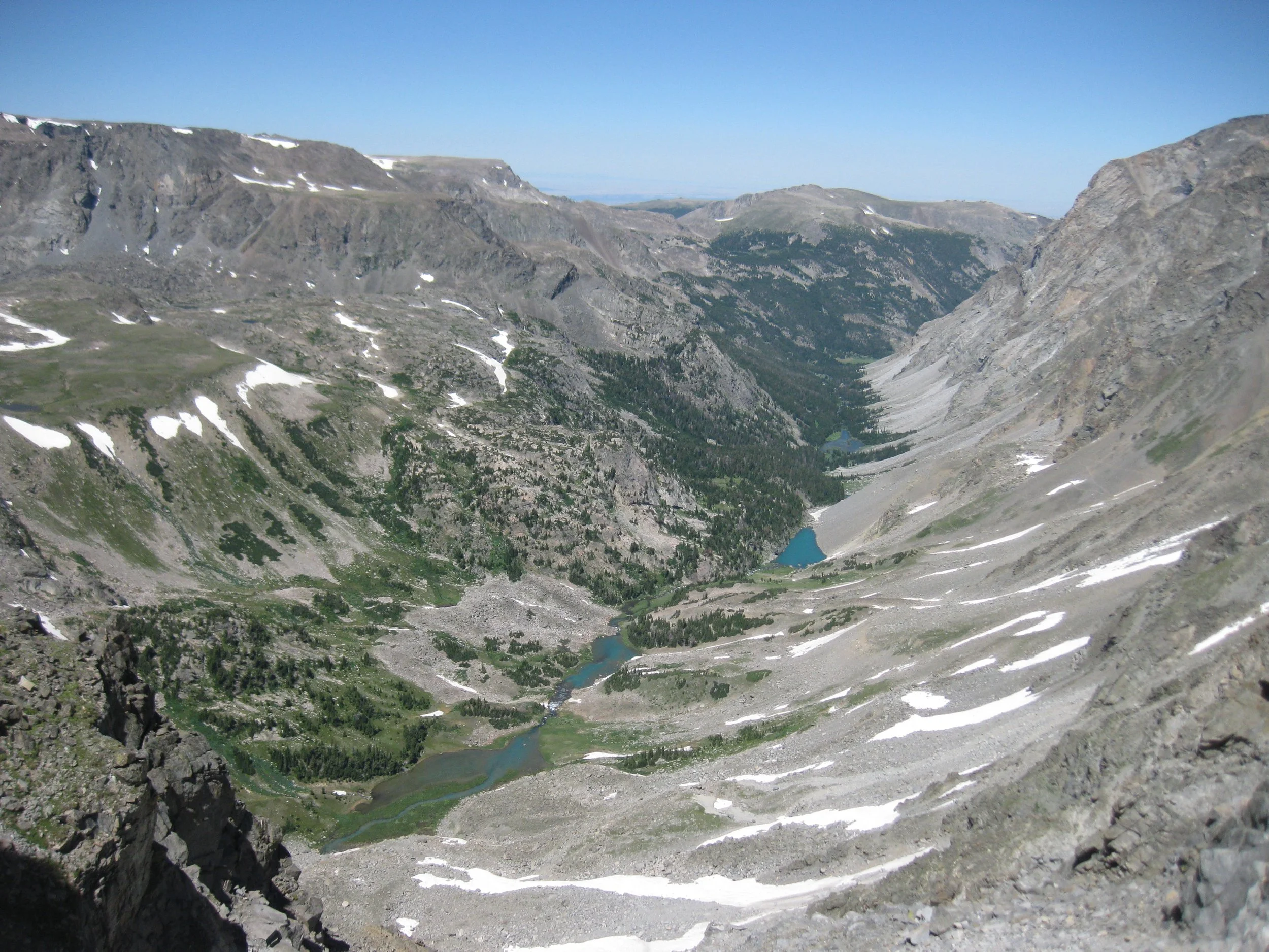 A panoramic view of a mountainous landscape with rocky terrain, patches of snow, green valleys, and several small lakes. The scene includes rugged peaks under a clear blue sky.