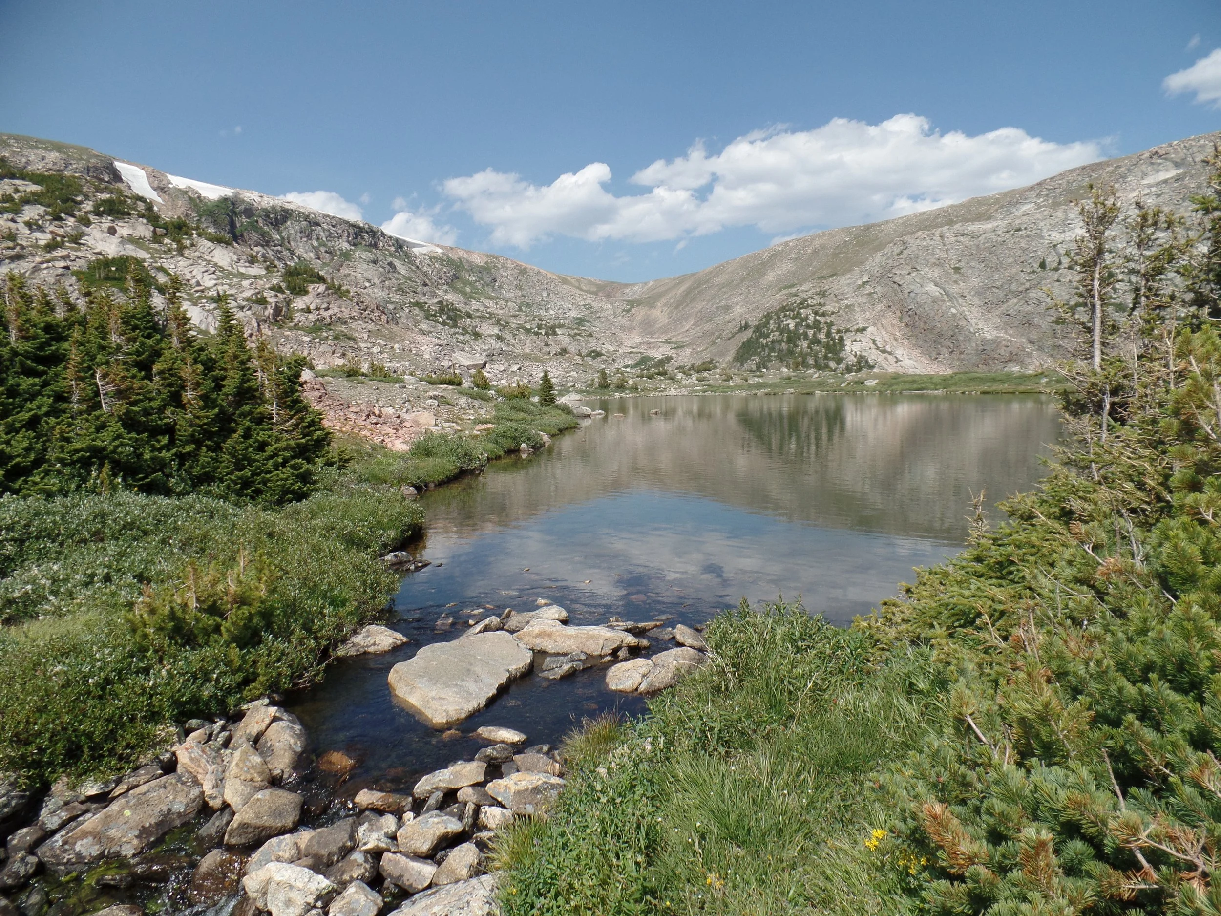Mountain landscape with a calm lake and rocky shore, surrounded by evergreen trees and rugged mountains under a blue sky with clouds.