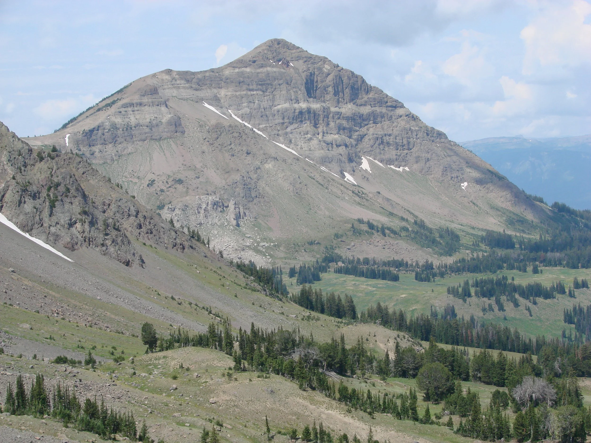 Mountain and valley landscape with sparse vegetation and patches of snow under a partly cloudy sky.