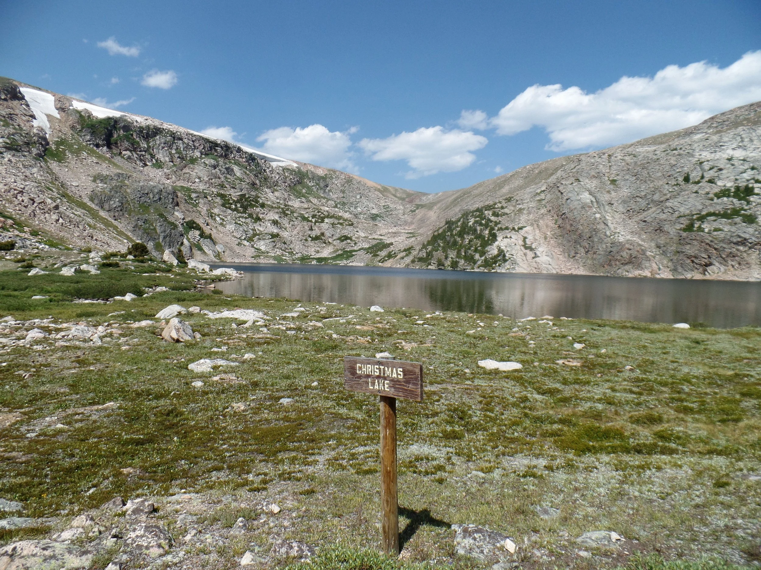 A tranquil mountain lake surrounded by rocky terrain and patches of grass under a clear blue sky. A wooden sign in the foreground reads 'Christmas Lake.'
